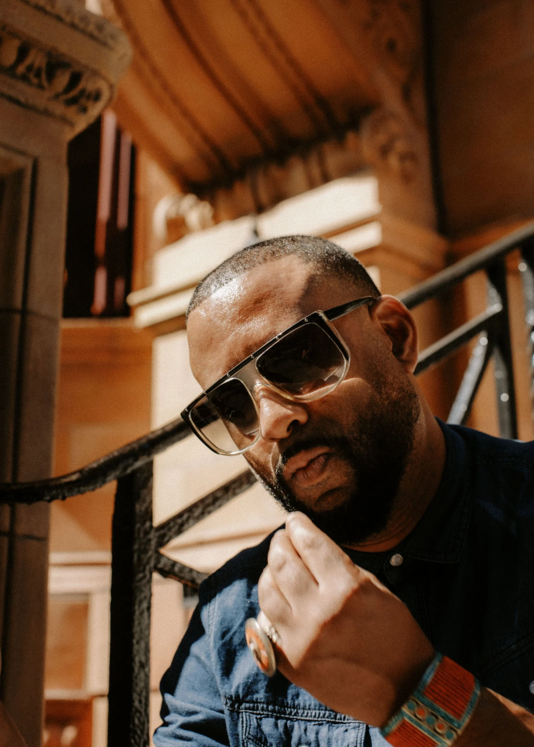 A man with a beard and sunglasses is sitting near a staircase railing, looking down. He is wearing a denim jacket and has a colorful woven bracelet on his wrist.