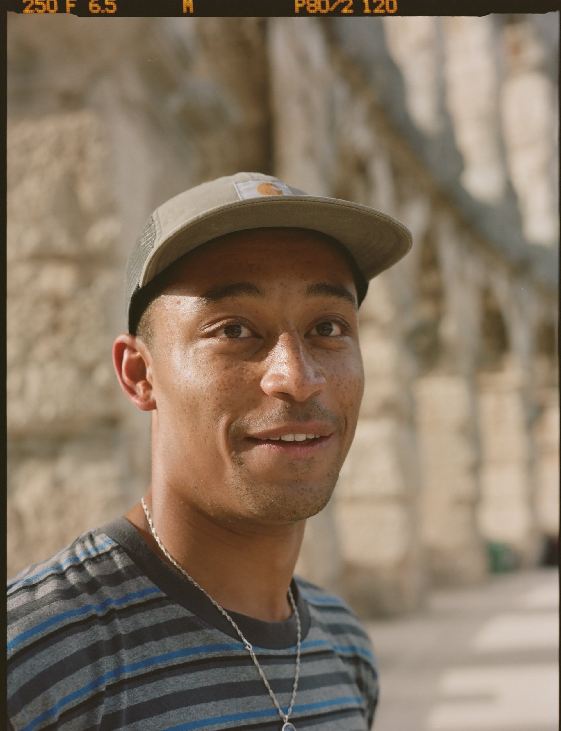 Close-up of a young man wearing a striped shirt, a baseball cap, and a chain necklace, standing outdoors against a blurred rocky background.