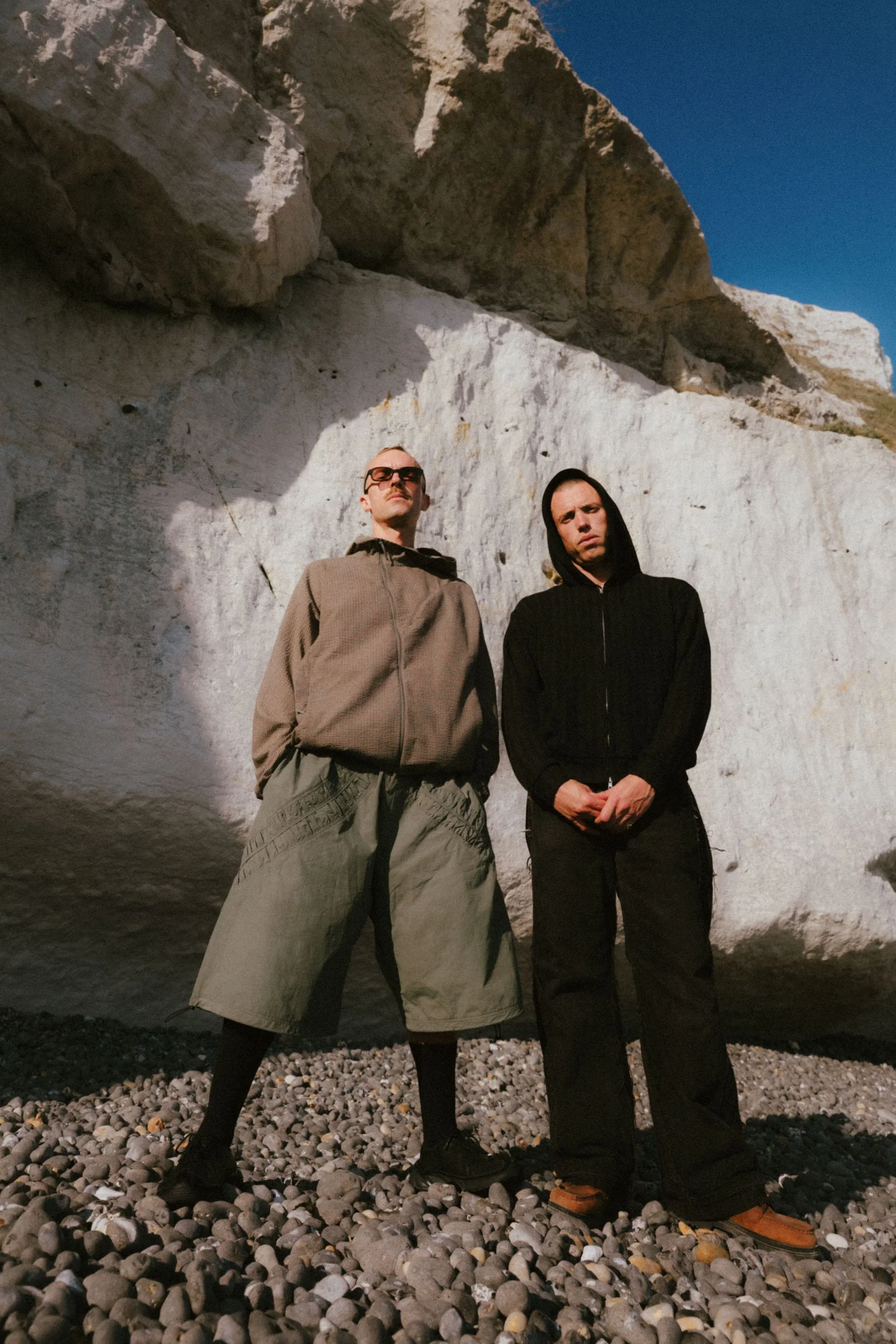 Two young men standing on a rocky beach in front of a white cliff with a clear blue sky above. One man wears sunglasses and a brown jacket, the other has a black hooded sweatshirt. Both have serious expressions.