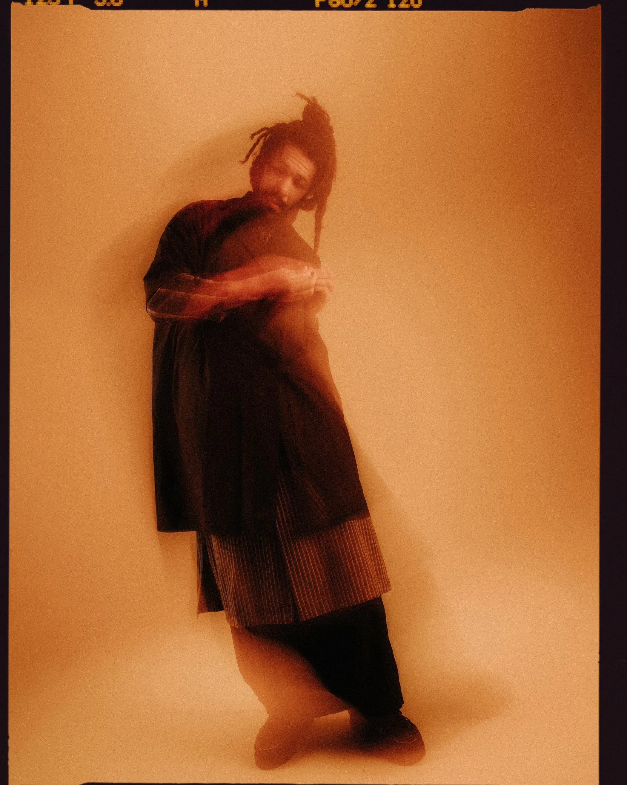 A person with dreadlocks and a beard standing against a plain wall, wearing a plaid shirt over a dark t-shirt and patterned pants, with a casual stance and looking at the camera.