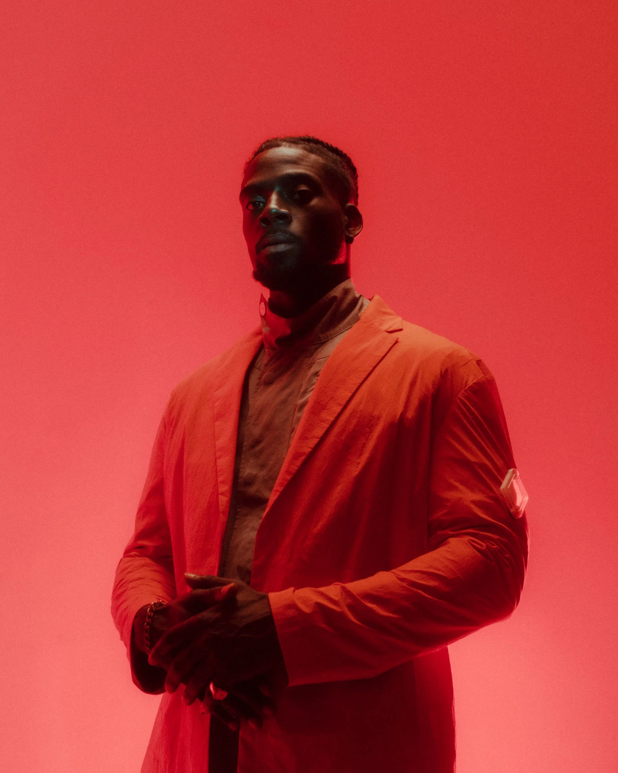 A man with dark skin, styled hair, and a beard, dressed in an orange blazer over a beige shirt, standing against a solid red background.