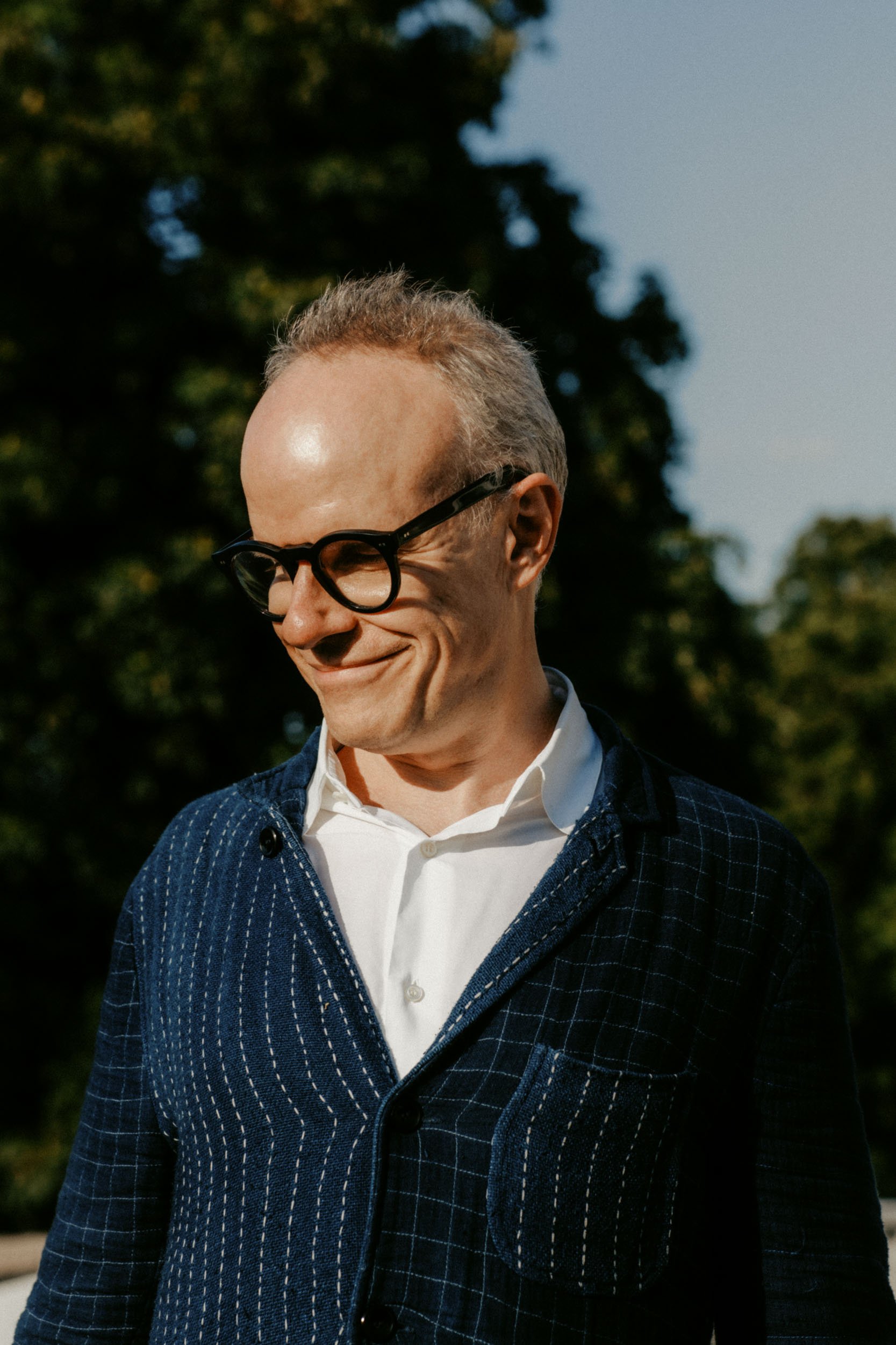 A man with glasses smiling outdoors during daytime, wearing a navy blue checkered blazer and a white shirt, with trees in the background.
