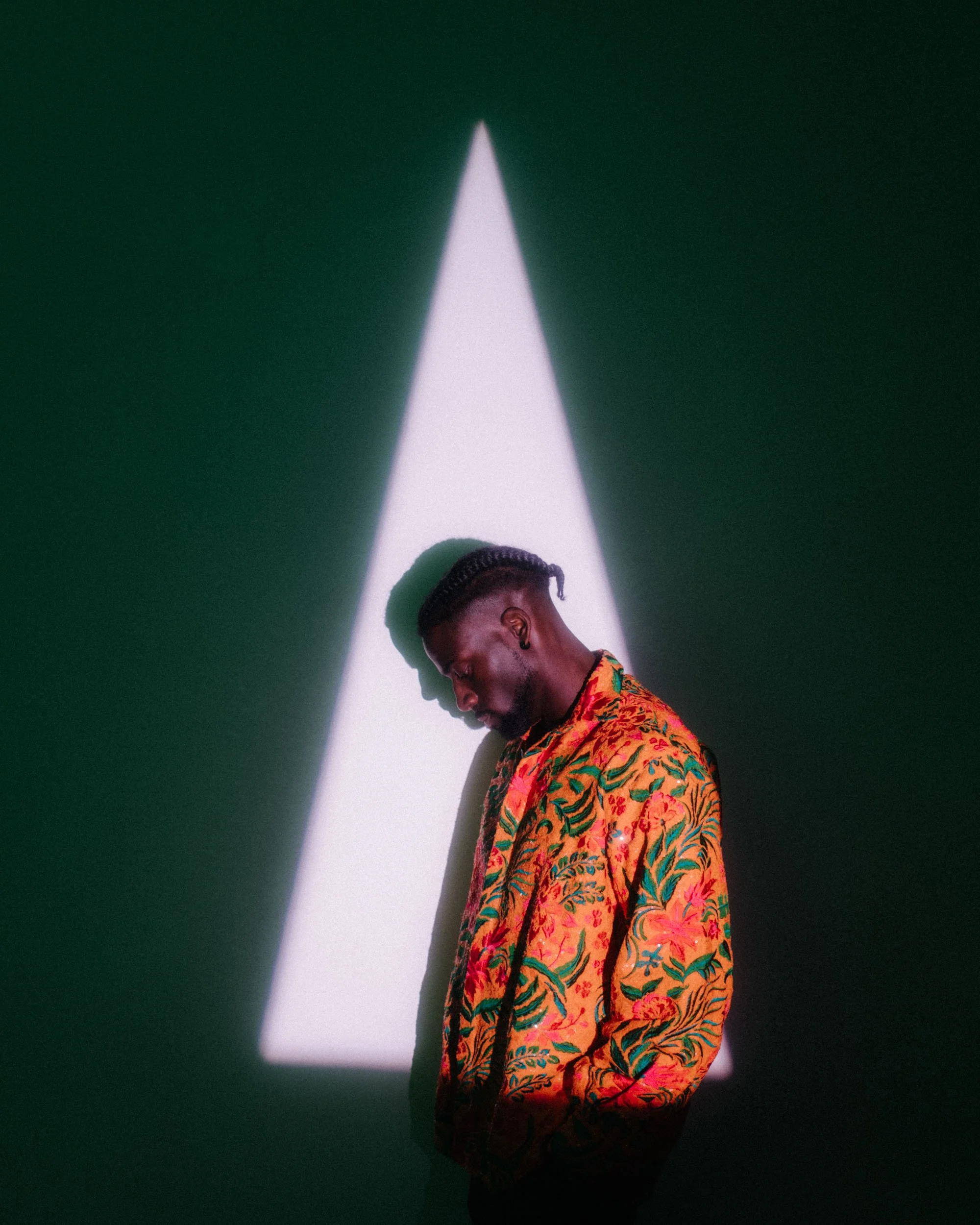 Young man with cornrows hairstyle wearing a vibrant, colorful patterned shirt standing in a shadowed space illuminated by a white triangular light from above.