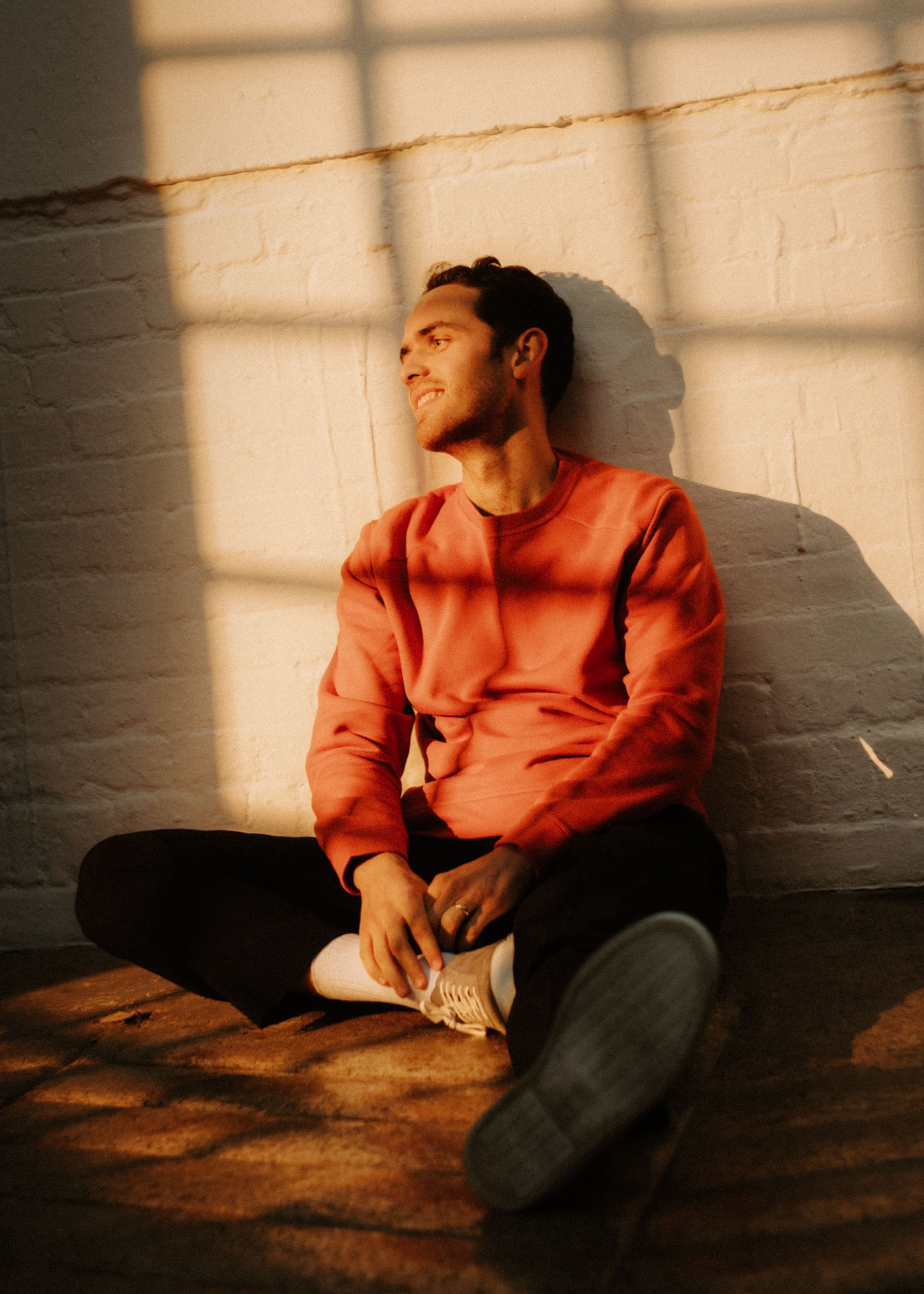 A young man sitting on the floor against a white brick wall, illuminated by warm sunlight filtering through window blinds, smiling and relaxed.