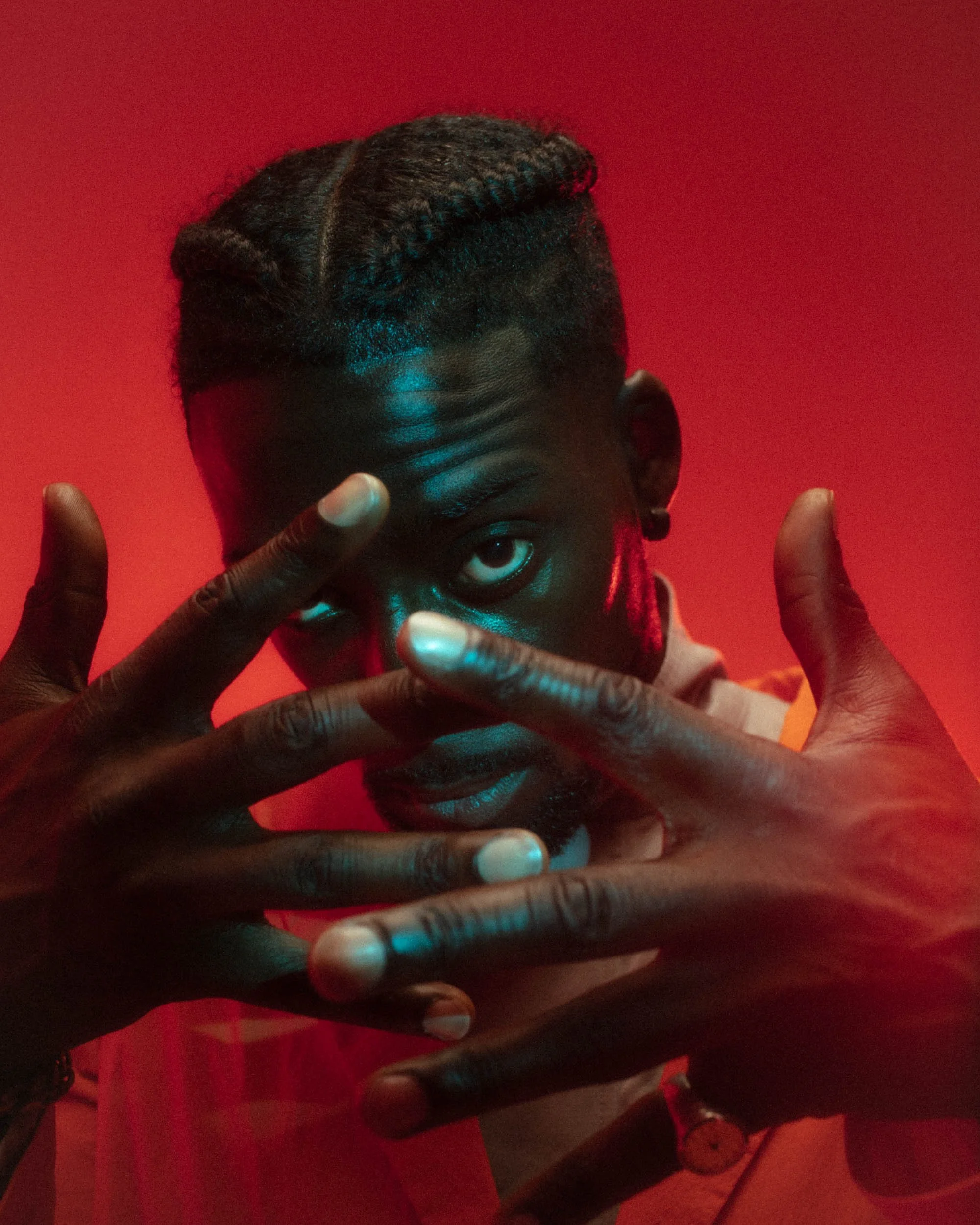 A person with dark skin and styled hair, making a peace sign with their fingers, against a red background.