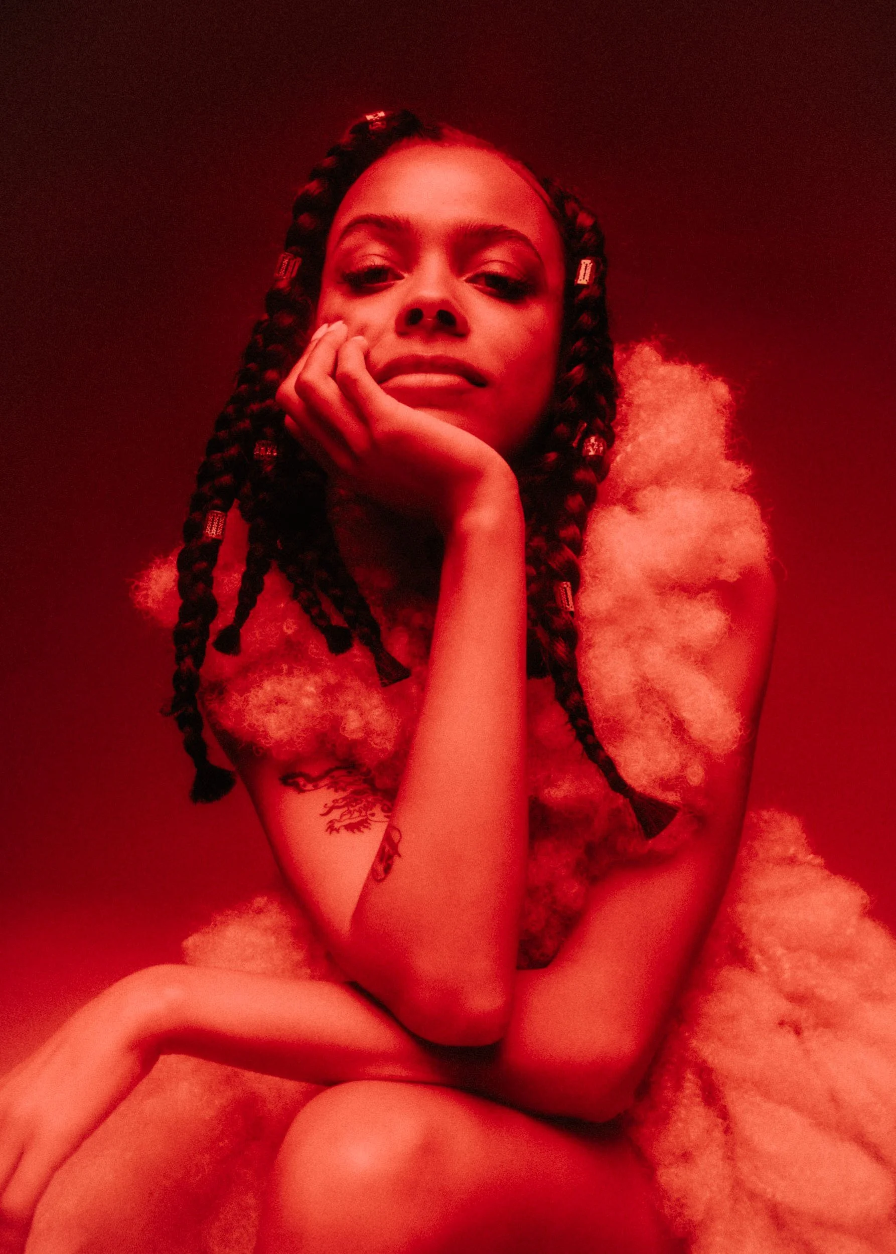 A young woman with braided hair and a tattoo on her arm, resting her chin on her hand, sitting on a plush, fluffy surface, with a red hue lighting the scene.