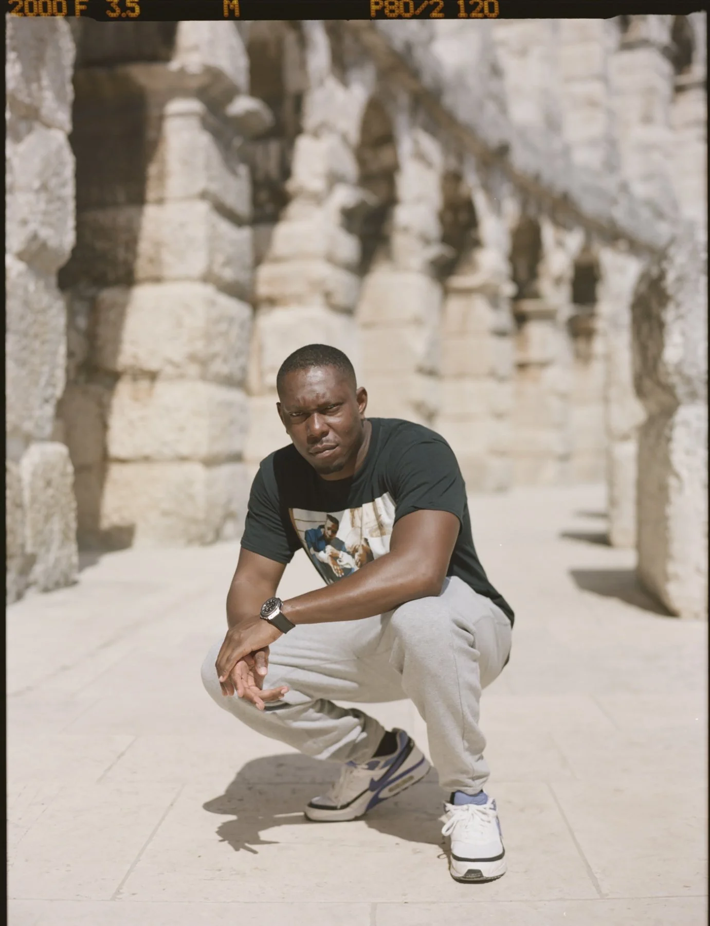 Dizzee Rascal crouching in front of ancient stone ruins, wearing a black T-shirt with a graphic, light gray pants, and sneakers, with a serious expression and a watch on his left wrist.