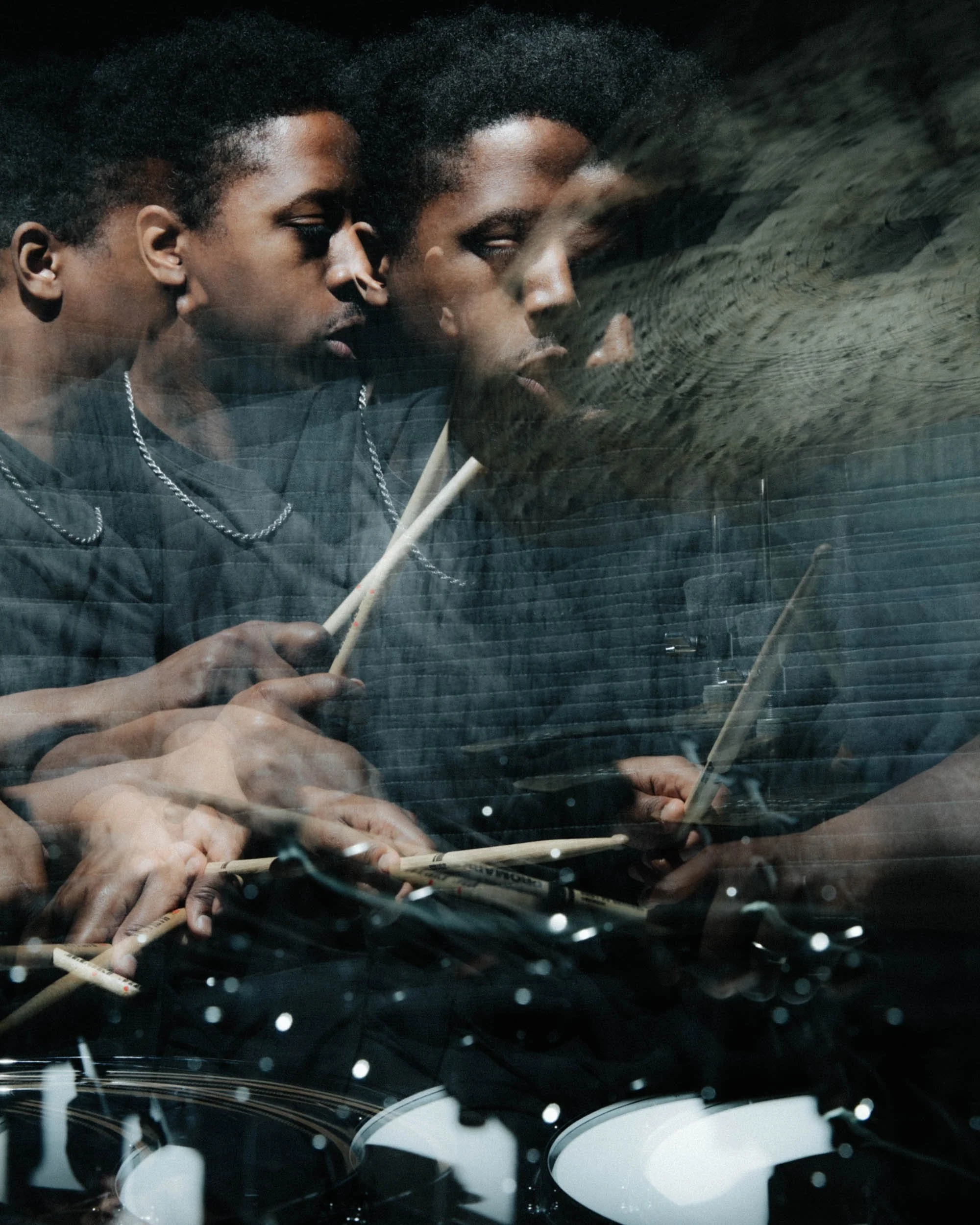 Reflection of two young men with short curly hair playing drums in a studio, with the view seen through a glass surface with water droplets.