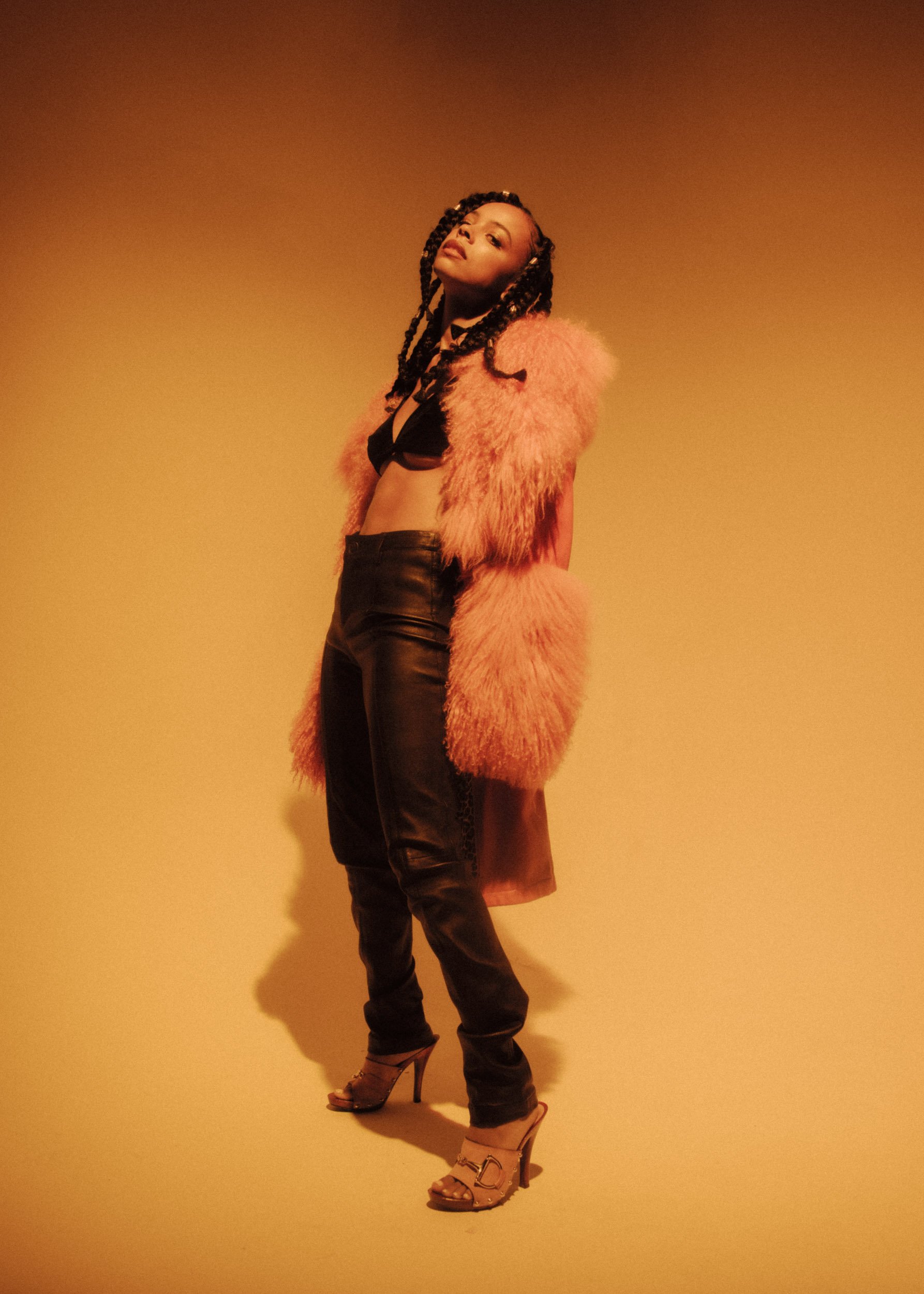 A woman posing against a plain beige wall, wearing a pink furry coat, a black crop top, high-waisted black pants, and beige high-heeled sandals.