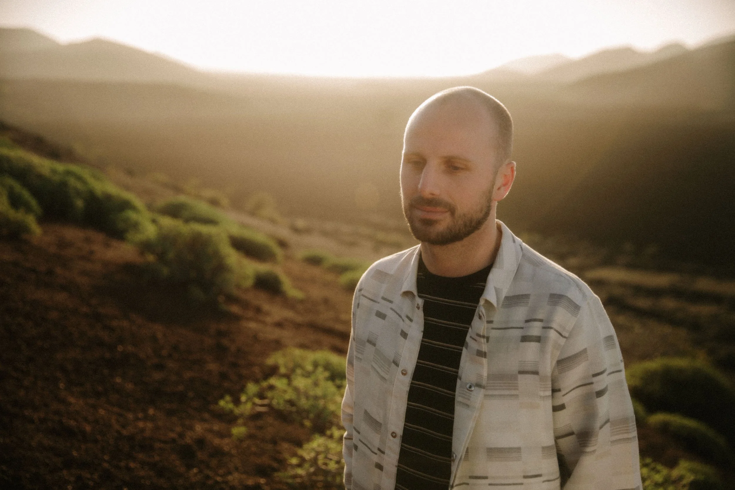 A man with a beard and short hair standing outdoors, wearing a striped shirt, with a hilly landscape and warm sunlight in the background.