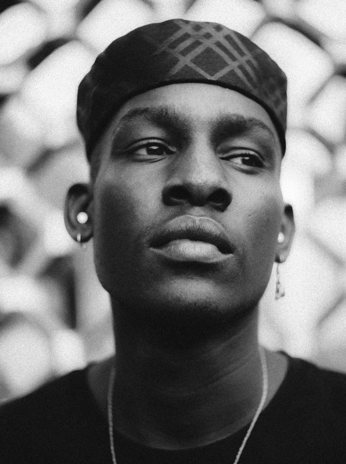A close-up black-and-white portrait of a person wearing a headscarf and earrings, with a blurred background.