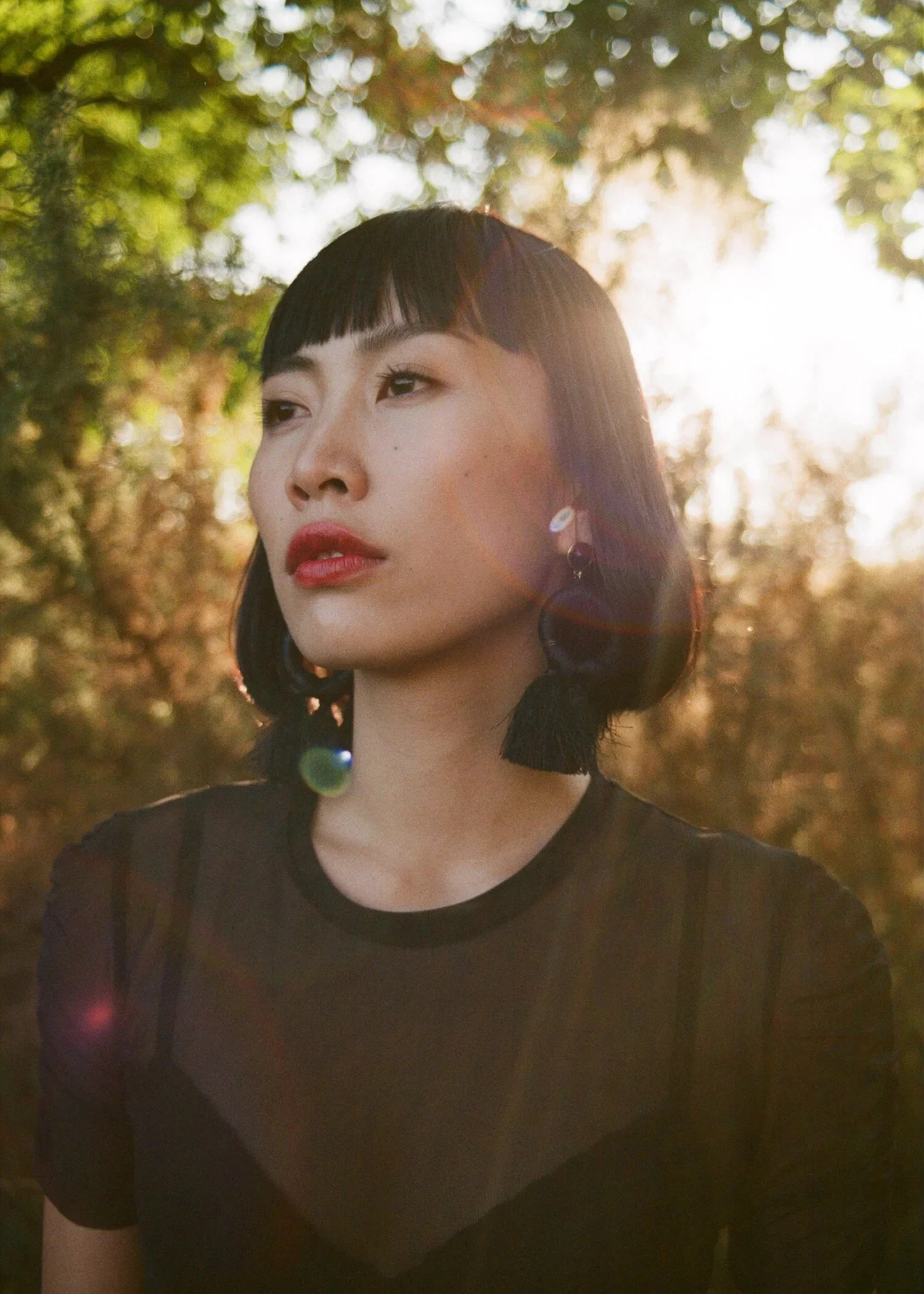 A young woman with dark hair and bangs, wearing earrings and a black top, is photographed outdoors with sunlight filtering through trees in the background.