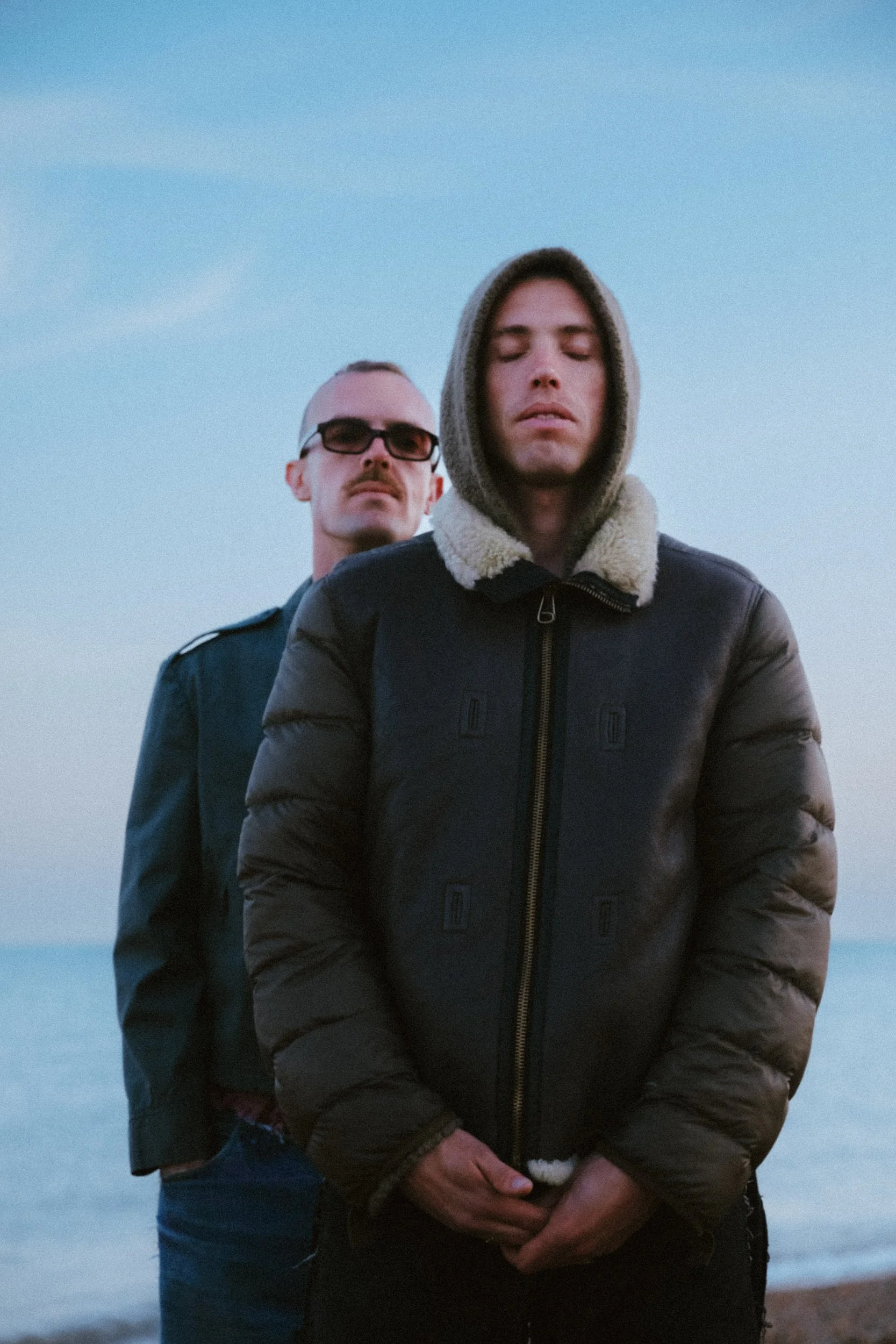 Two men standing outdoors near the water, one in a hoodie and the other in a jacket with a shearling collar, both with their eyes closed.