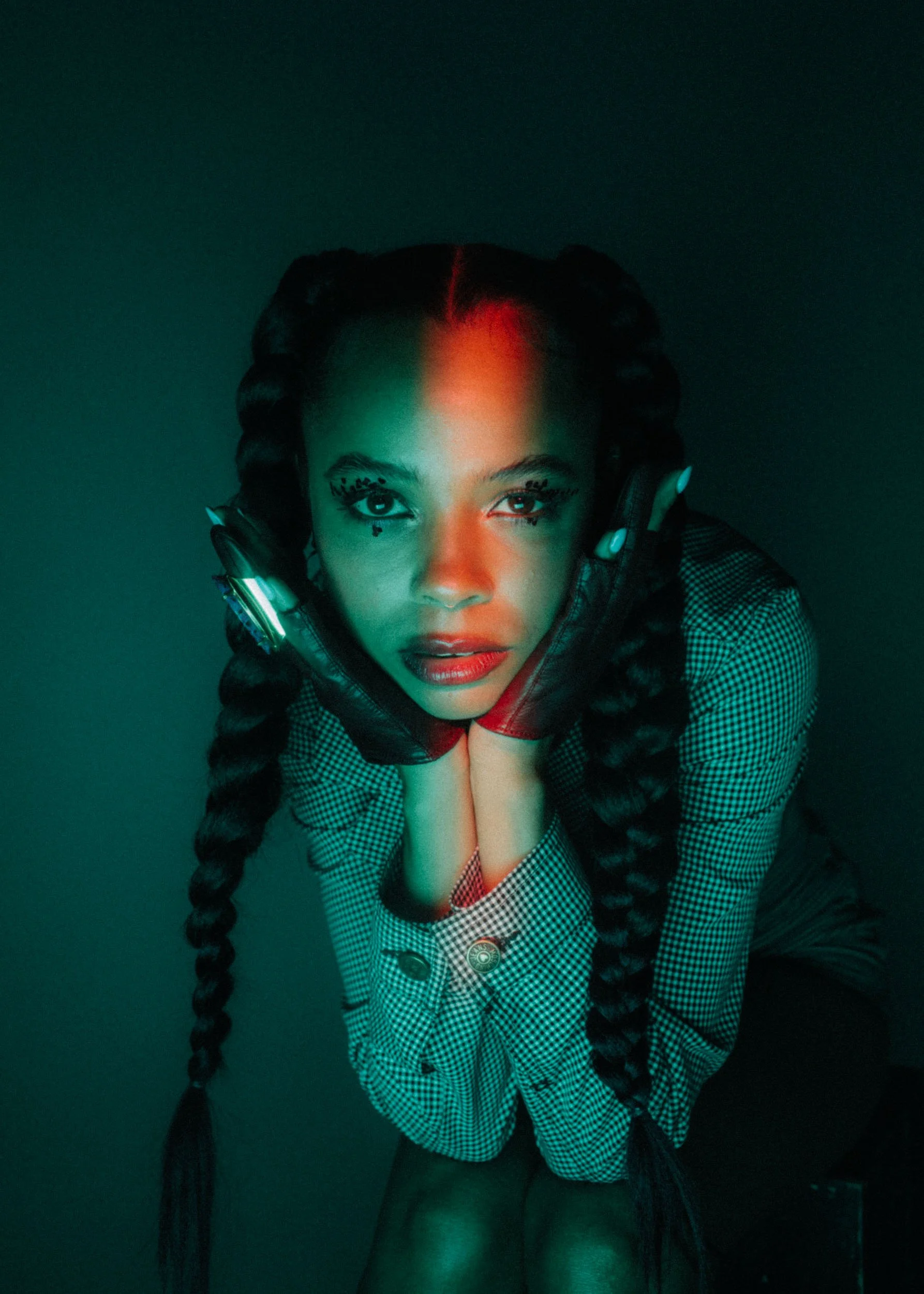A young woman with braided hair, wearing headphones, a black and white checkered shirt, and black gloves, is looking directly at the camera with a serious expression, against a dark background.