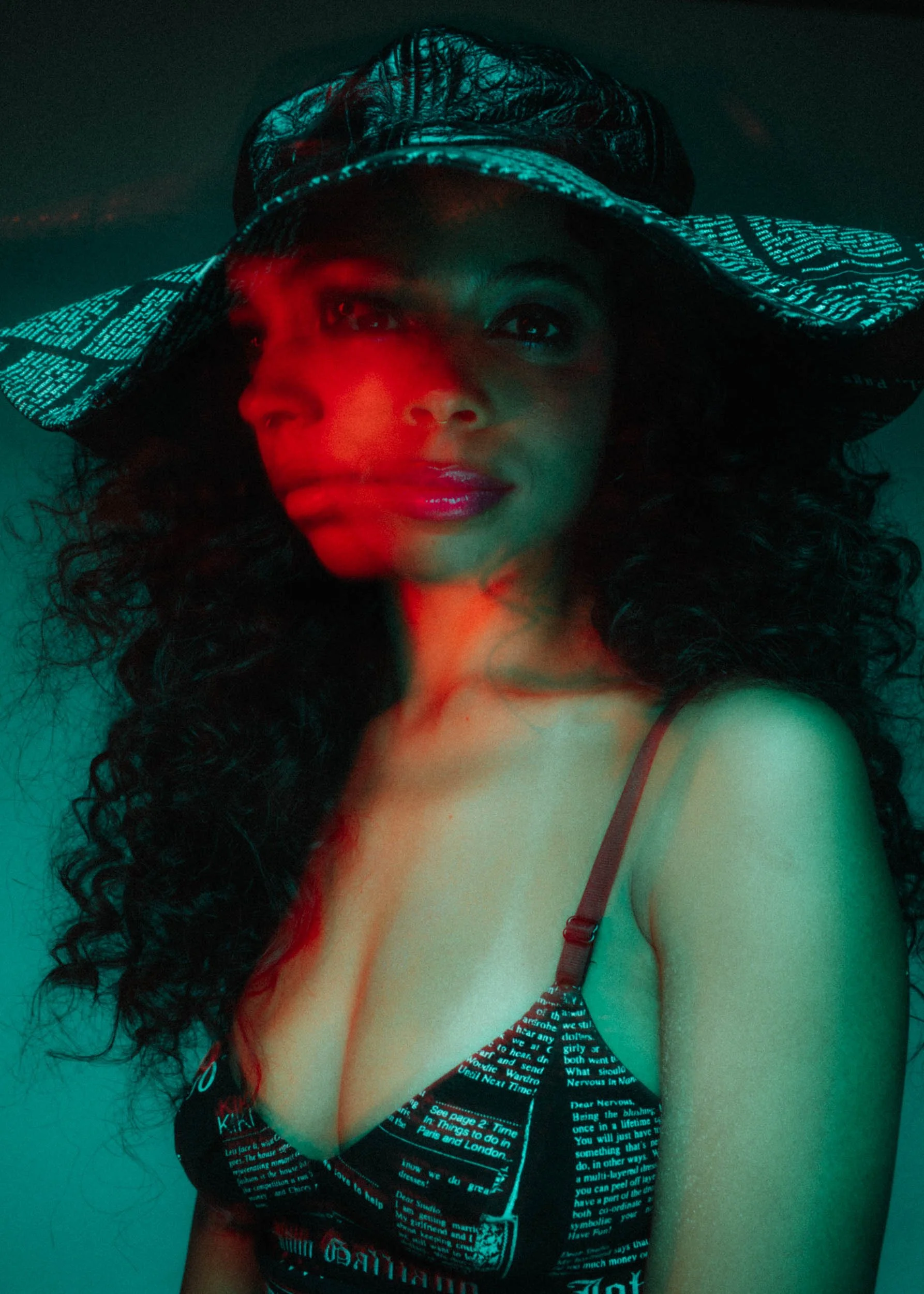 Close-up of a woman with curly hair wearing a black and white text-printed wide-brimmed hat and a matching dress with text pattern, illuminated by red and teal lighting.