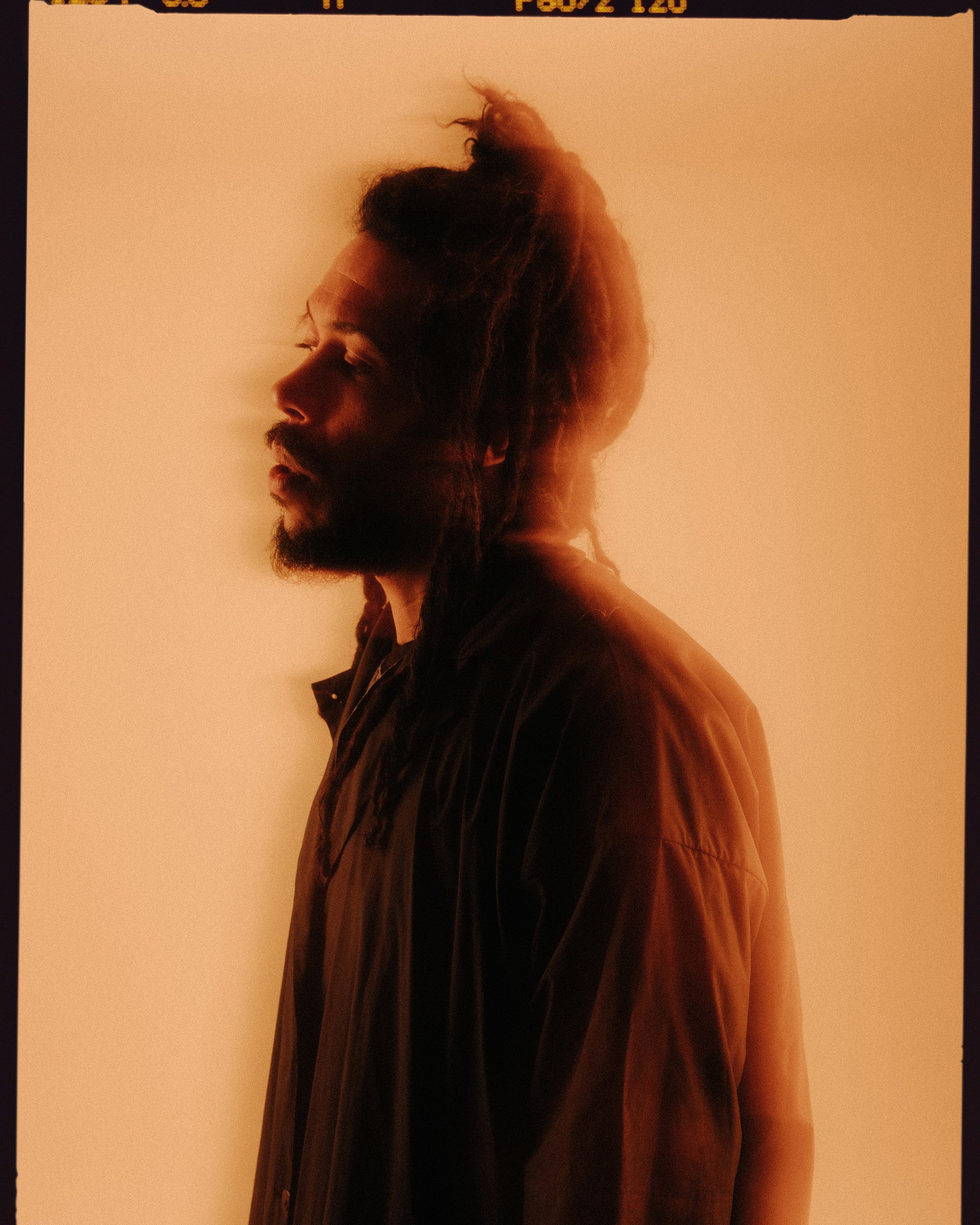 Side profile of a man with a beard and dreadlocks, wearing a brown jacket, standing against a beige background, with warm lighting.