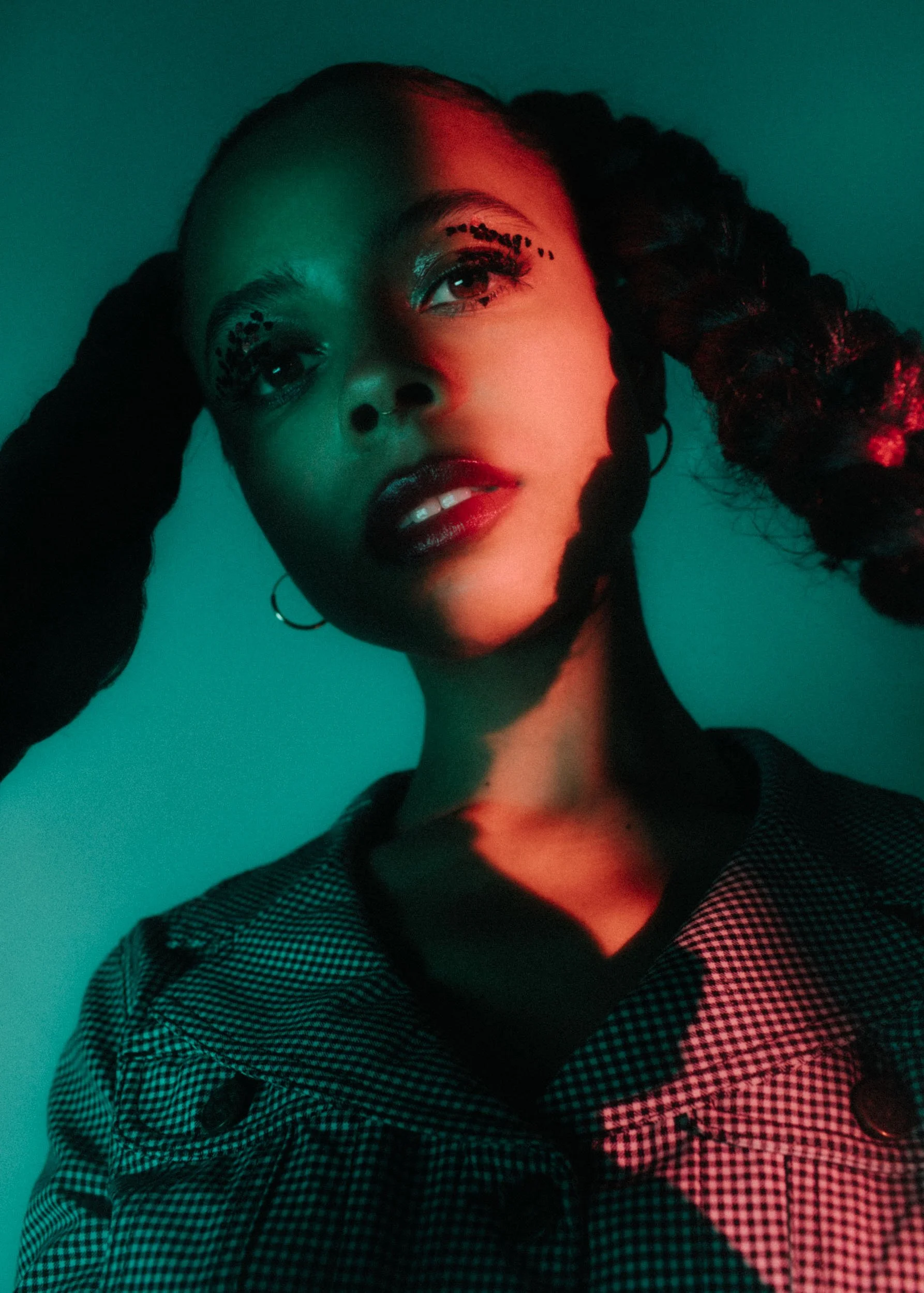 A young woman with curly hair styled in braids, wearing makeup with bold eye makeup and dark lipstick, dressed in a black and white checkered shirt, positioned against a teal background with red and green lighting.