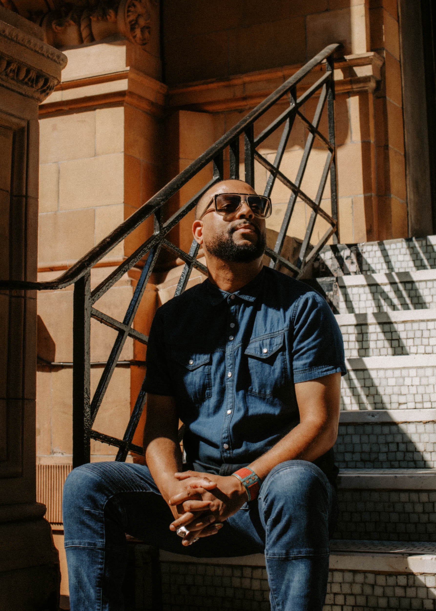 A man with a beard and sunglasses sitting on stairs, wearing a denim shirt and jeans, with arms resting on his laps, in a dimly lit area with brick and wood decor.