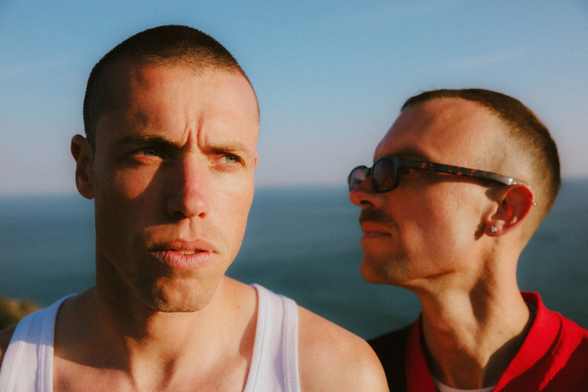Close-up of two young men with buzz cuts, one wearing sunglasses, standing outdoors against a bright blue sky.