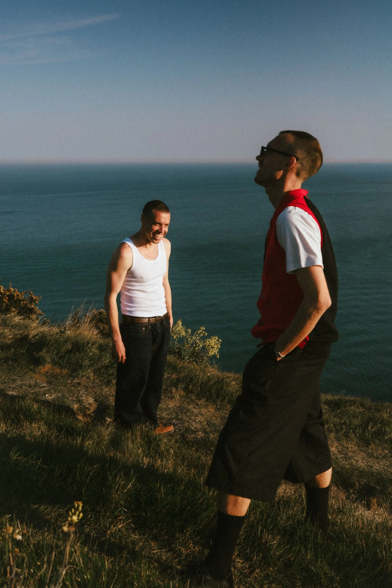 Two men are standing outdoors on a grassy hill near the ocean, laughing and talking during sunset.