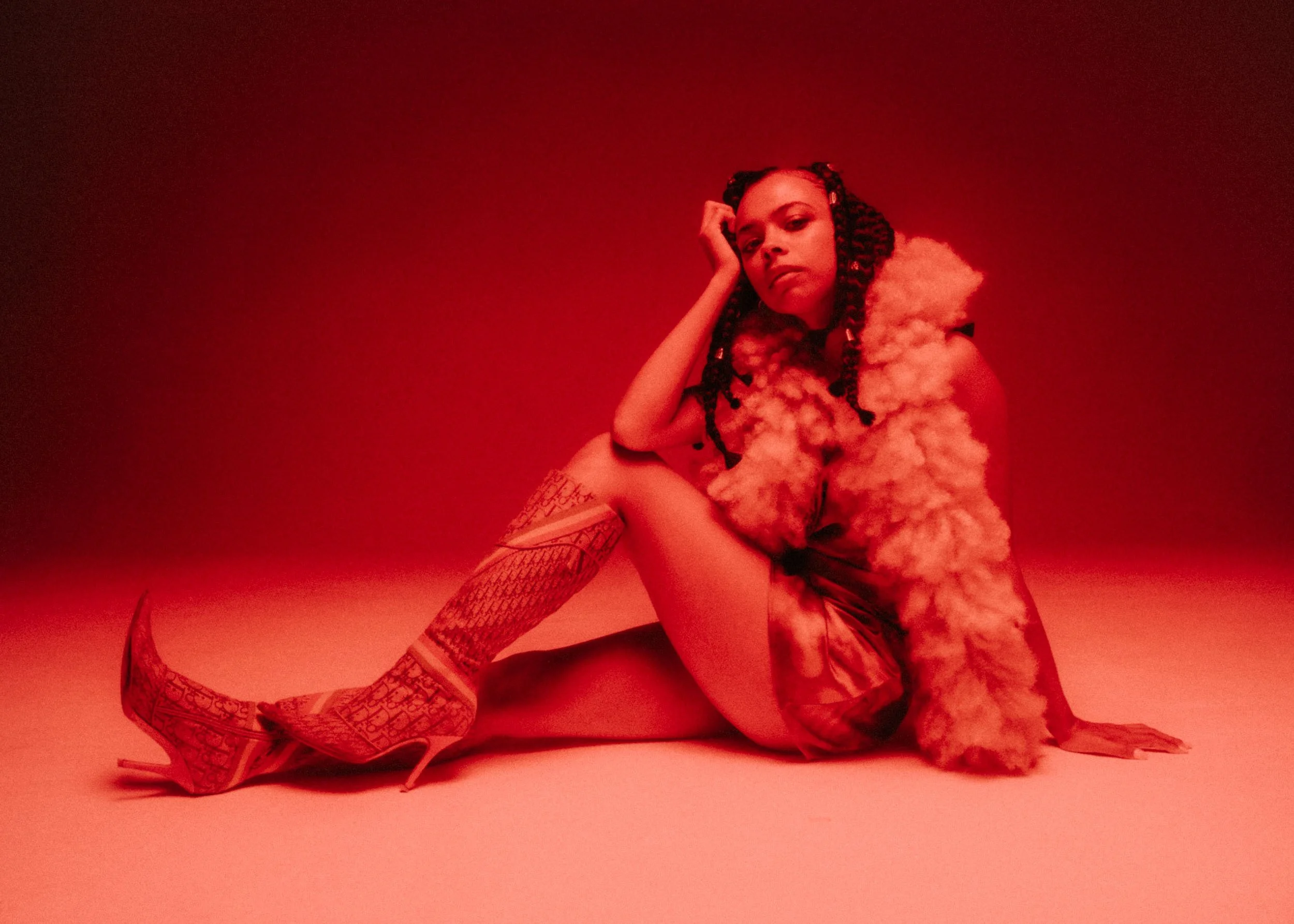 A woman with long braids sitting on the floor with her head resting on her hand, wearing high heels, fishnet stockings, and a fluffy coat, against a red background.