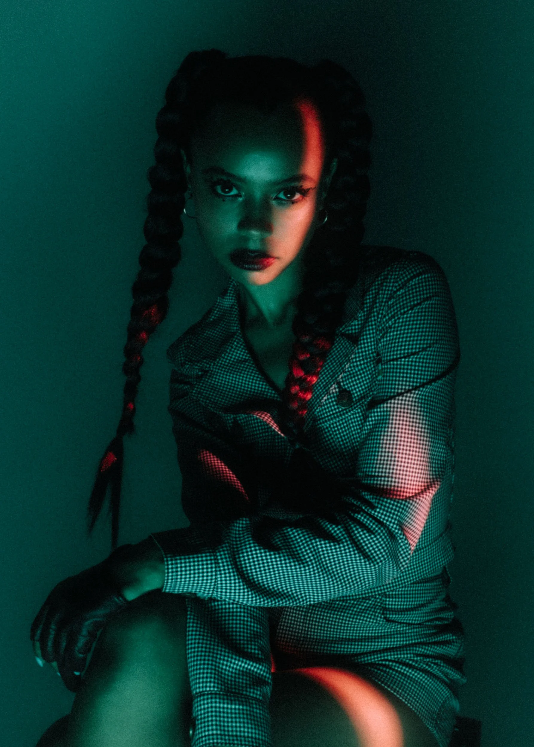 A woman with dark braided hair and dark lipstick, wearing a checkered blazer, looks directly at the camera with an intense gaze, illuminated by green lighting.