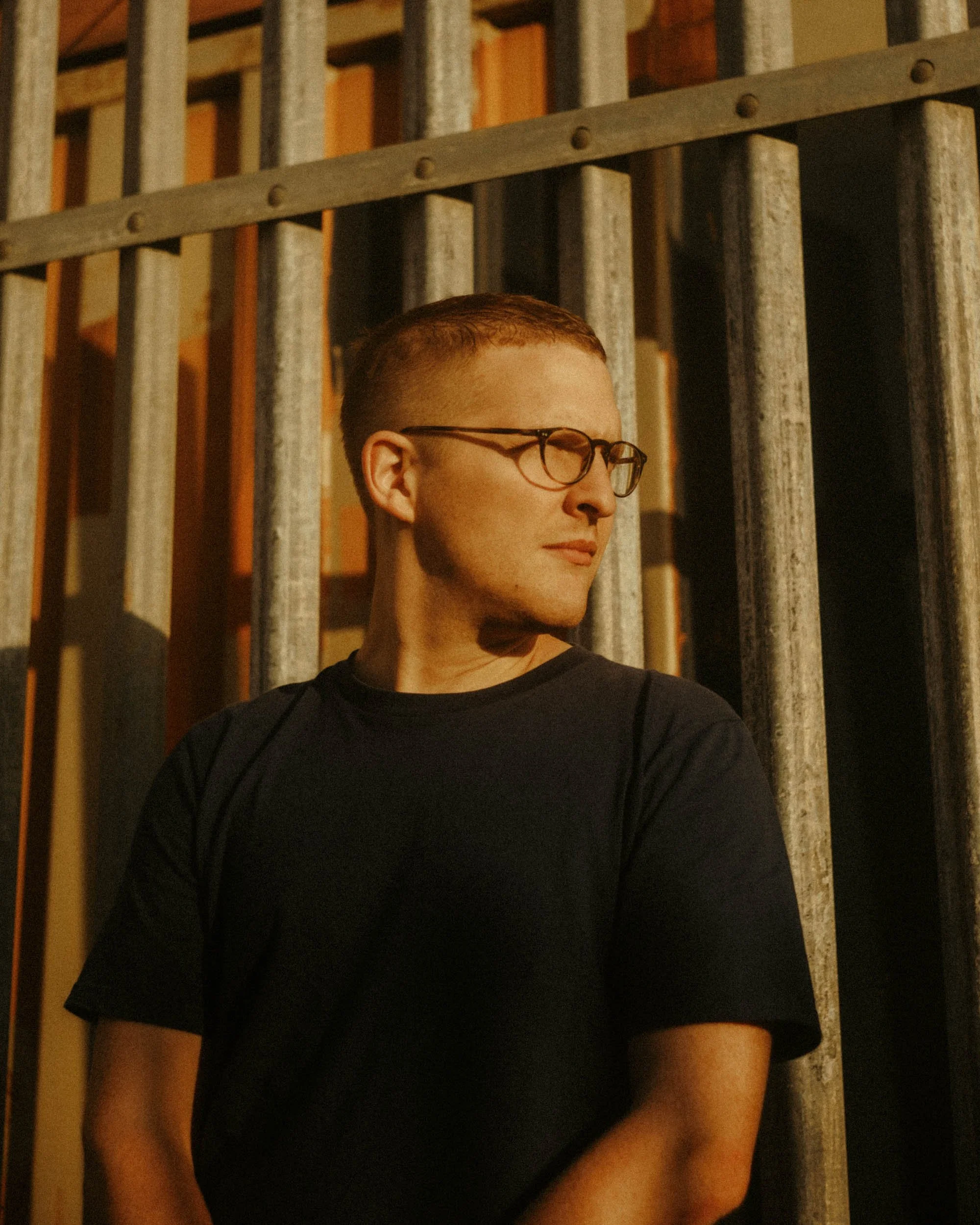 A person with short hair and glasses standing in front of a metal and wooden fence, looking to the side.
