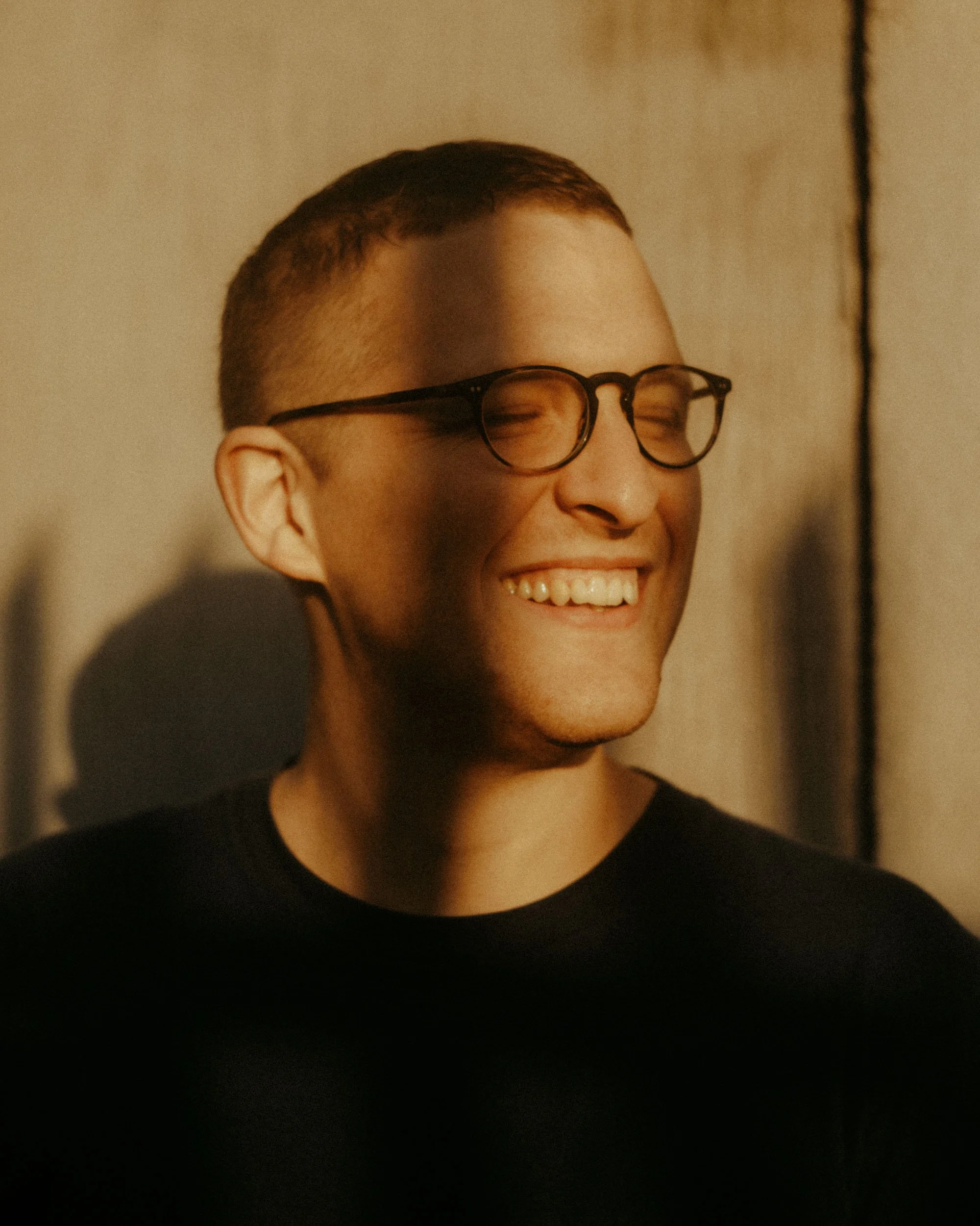 A person with short hair, wearing glasses, smiling with closed eyes, and wearing a black shirt, against a plain wall.