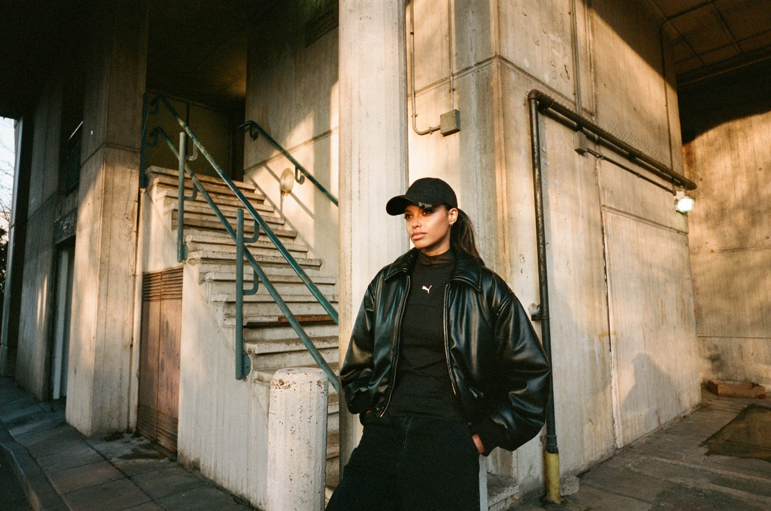 Woman wearing a black baseball cap and leather jacket standing next to a concrete building exterior with stairs and pipes.