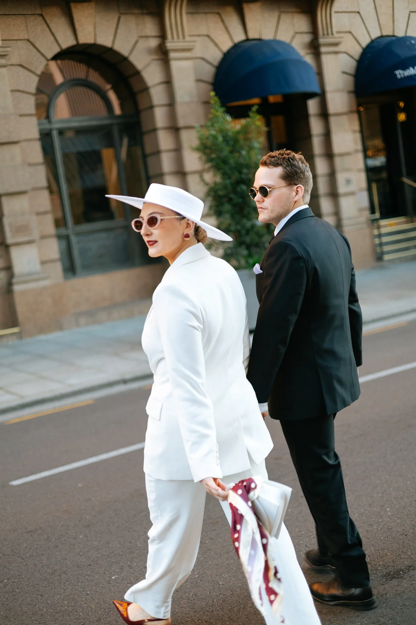 A woman in a stylish white suit with a large white hat and round sunglasses walking with a man in a black suit and sunglasses on a city street with historic buildings and arched windows in the background.