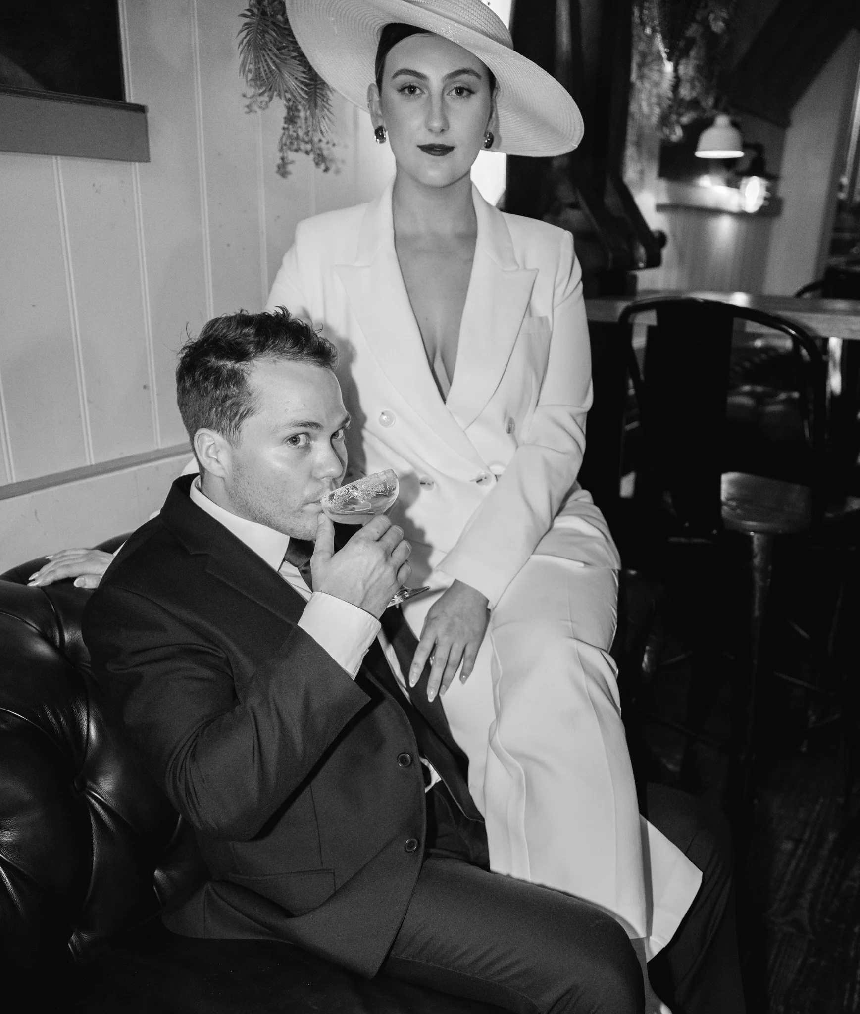 Black and white photo of a woman in elegant attire, wearing a wide-brimmed hat, sitting next to a man in a tuxedo holding a drink and looking intently at the camera, in a stylish indoor setting.