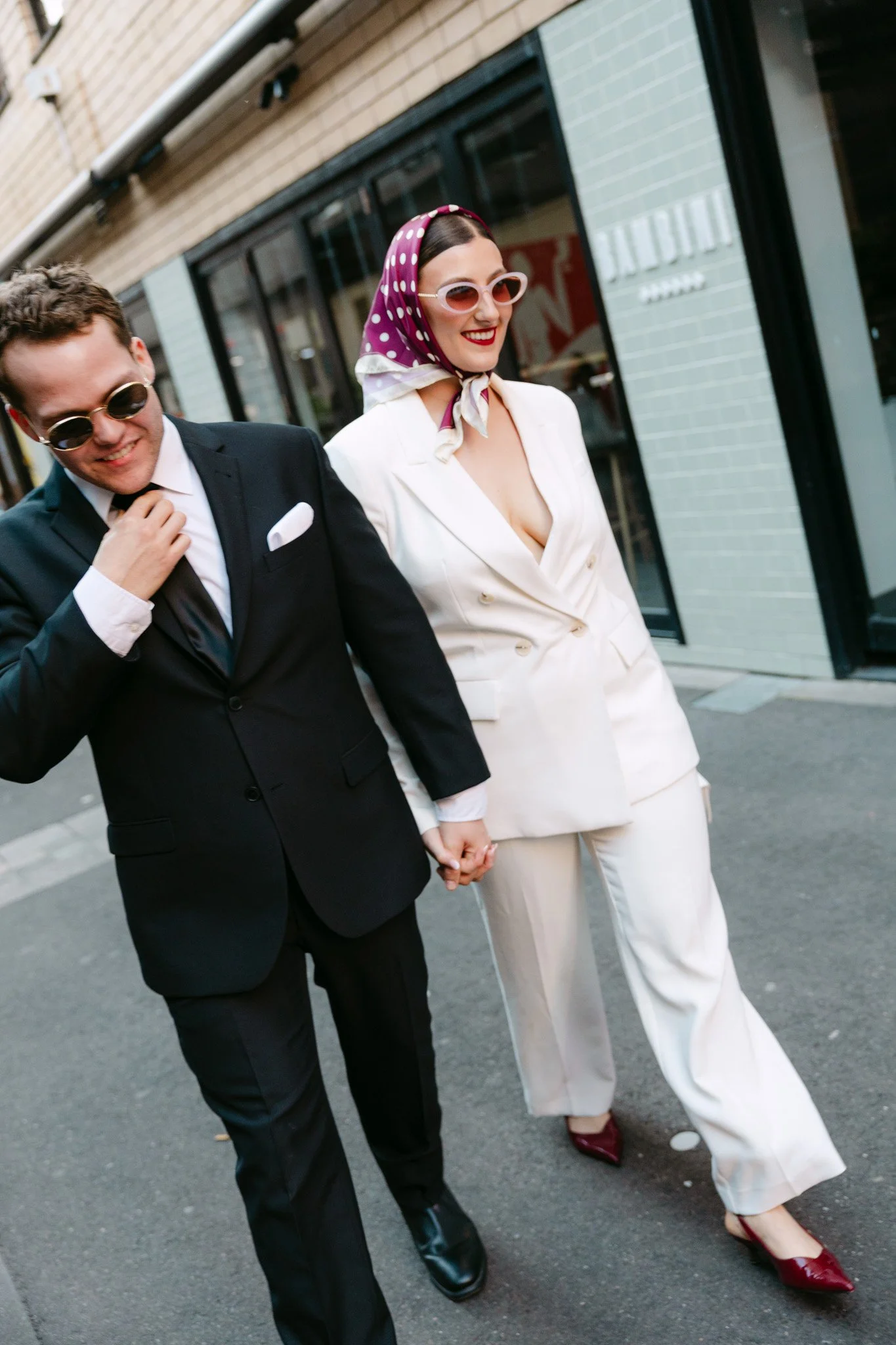 A couple walking hand in hand on a city street, dressed in vintage-style clothing, with the woman wearing a purple polka dot headscarf, white blazer, and sunglasses, and the man in a black suit and sunglasses.