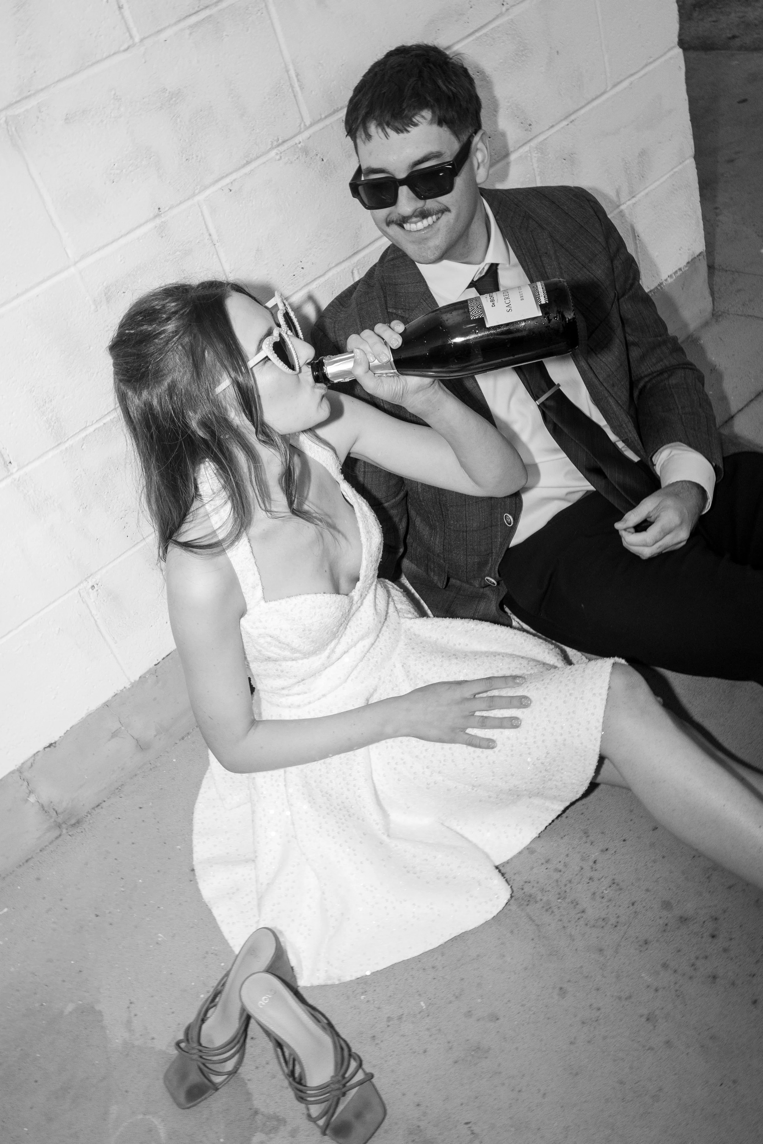 A woman in a white dress sitting on the ground, drinking from a bottle held by a man in a suit and sunglasses, with her hand on her lap. Their shoes are on the ground nearby.