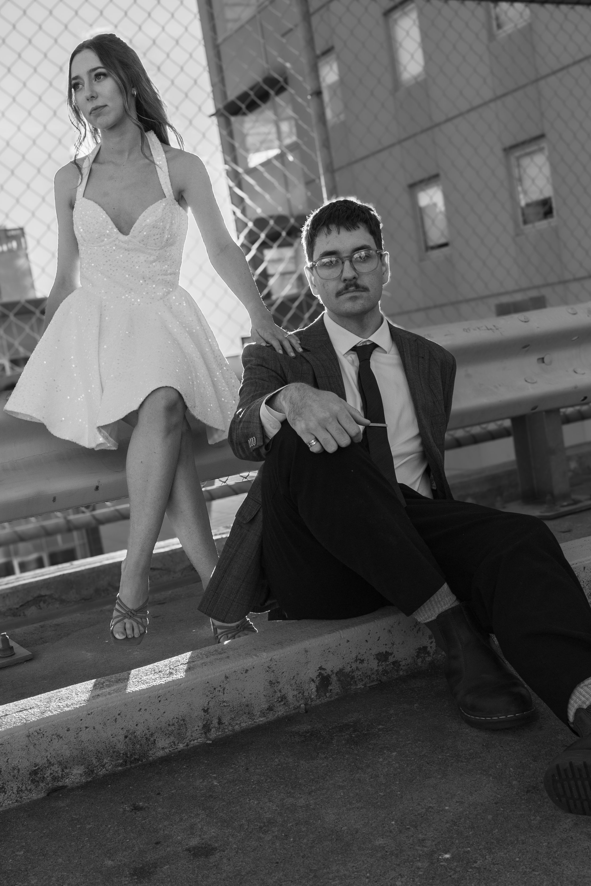 A black and white photo of a woman in a dress with halter straps and high heels standing on a rooftop or bridge with a chain-link fence behind her. A man with glasses and a mustache in a suit is sitting on the ground, leaning against a bench or ledge, holding a cigarette.