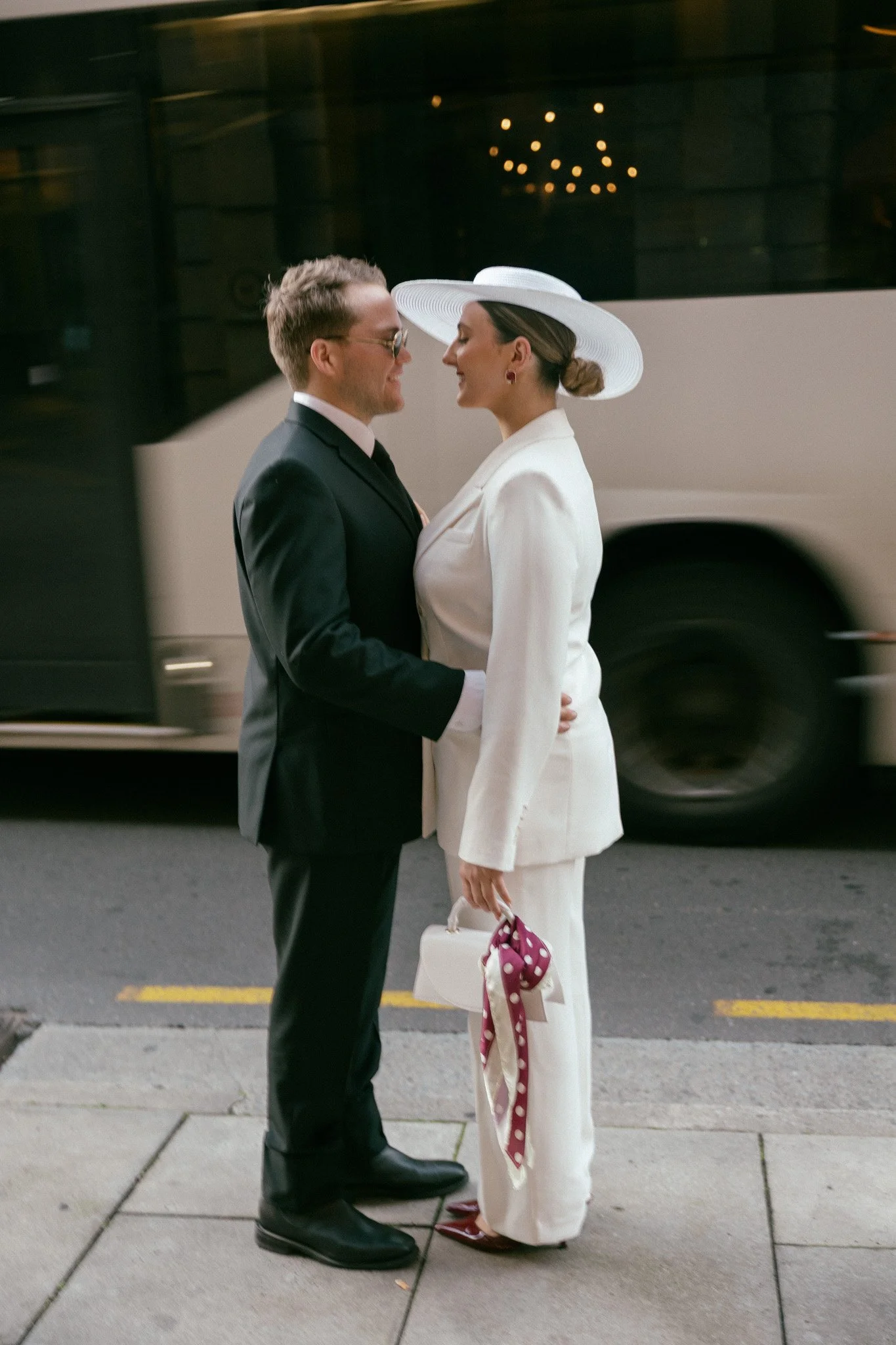 A man and woman dressed in formal attire, standing close and smiling to each other on a city sidewalk at night, with a bus passing behind them.