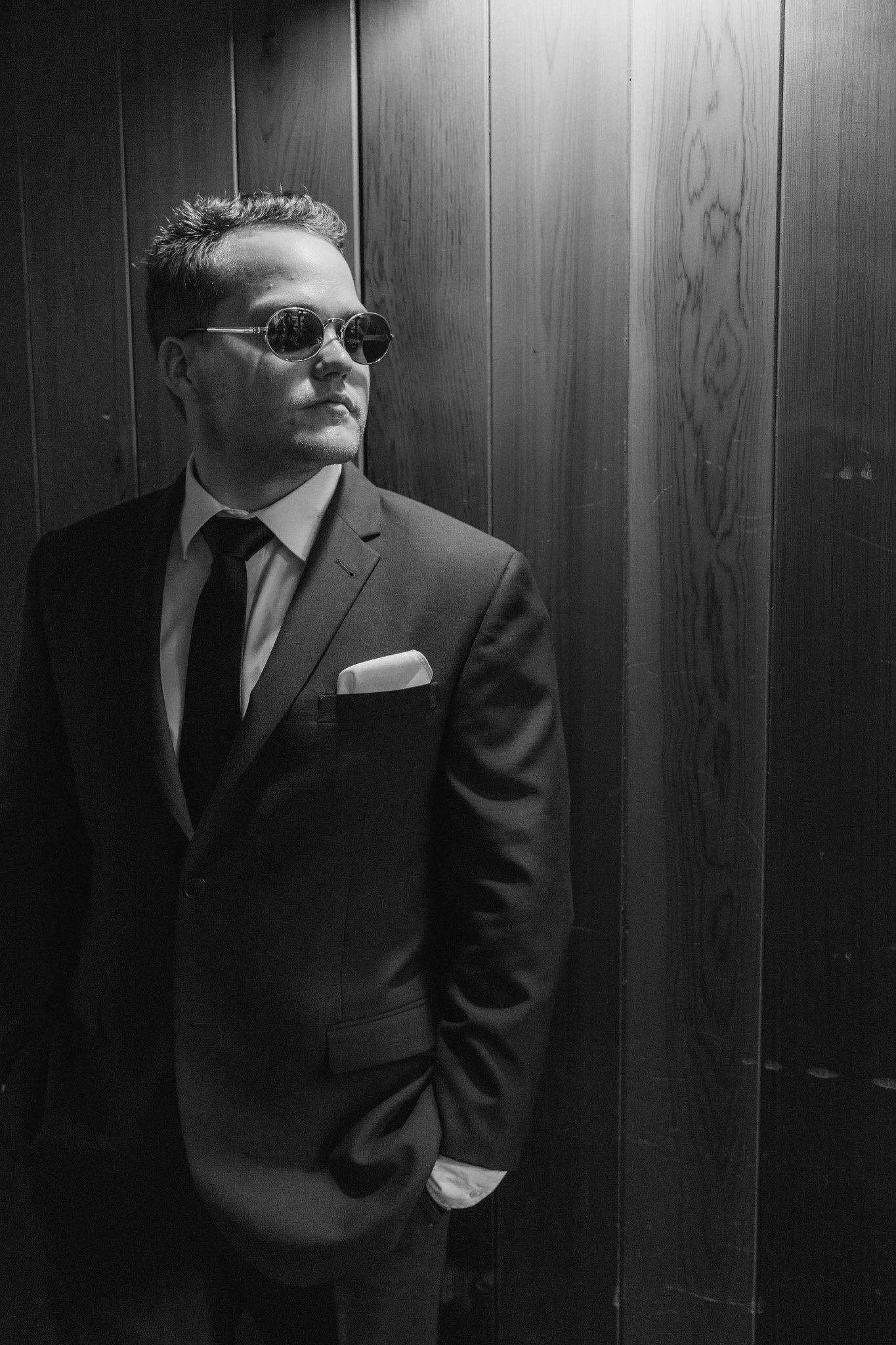 A man in a suit and sunglasses standing in an elevator with wood-paneled walls.