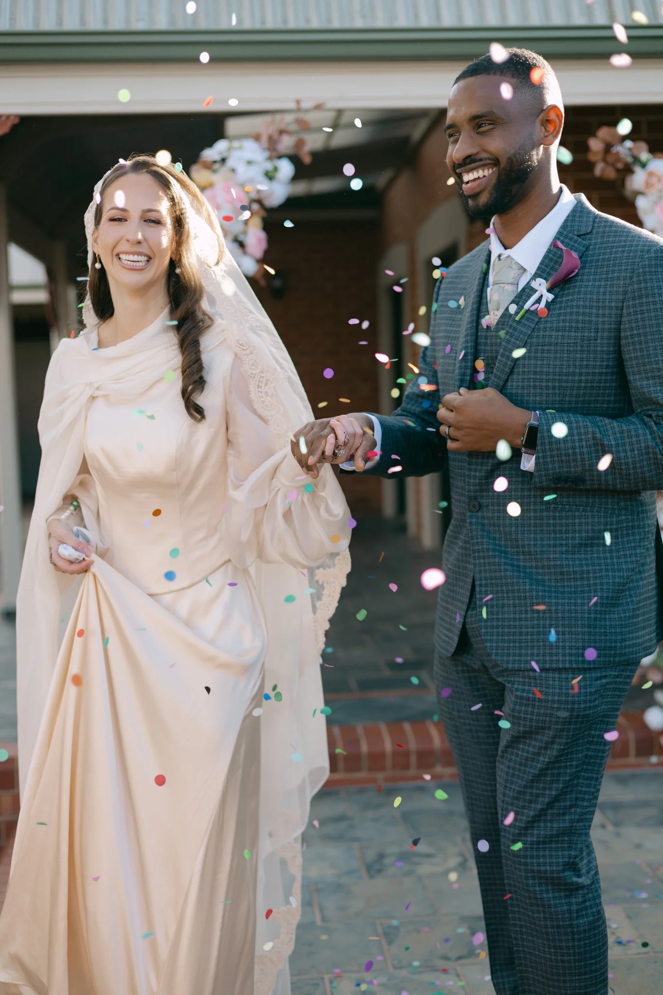 A bride and groom at their wedding celebration, smiling and holding hands, with colorful confetti falling around them, outside a building with floral decorations.