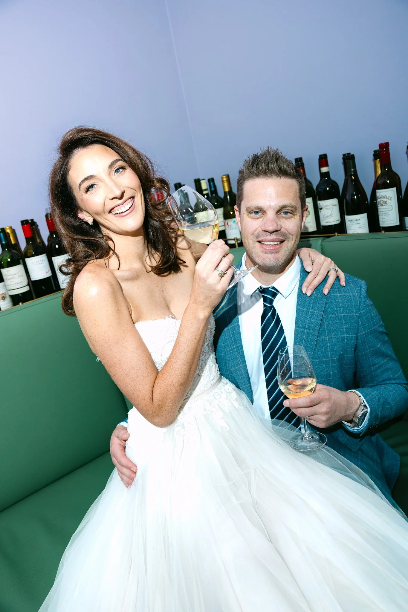 A smiling bride and groom at their wedding reception, holding glasses of white wine, with a backdrop of wine bottles on a shelf.