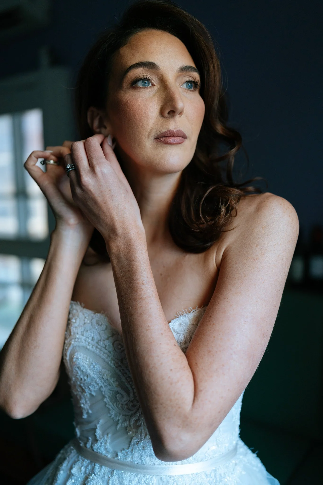 Close-up of a woman with blue eyes and dark wavy hair, wearing a strapless white lace wedding dress, adjusting her earring, with a dark background and a window reflecting in the distance.