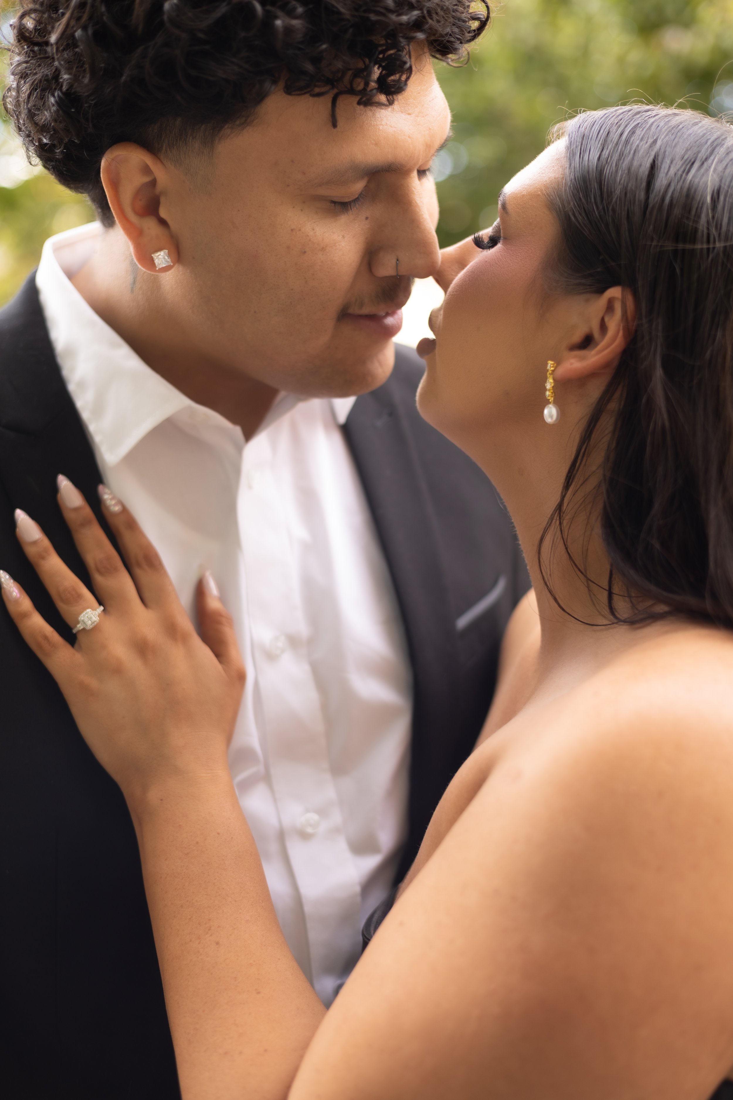 A couple is close together, about to kiss outdoors with a blurred green background. The man has curly hair, a nose piercing, and is wearing a black suit with a white shirt. The woman has long dark hair, gold earrings with pearls, and is topless.