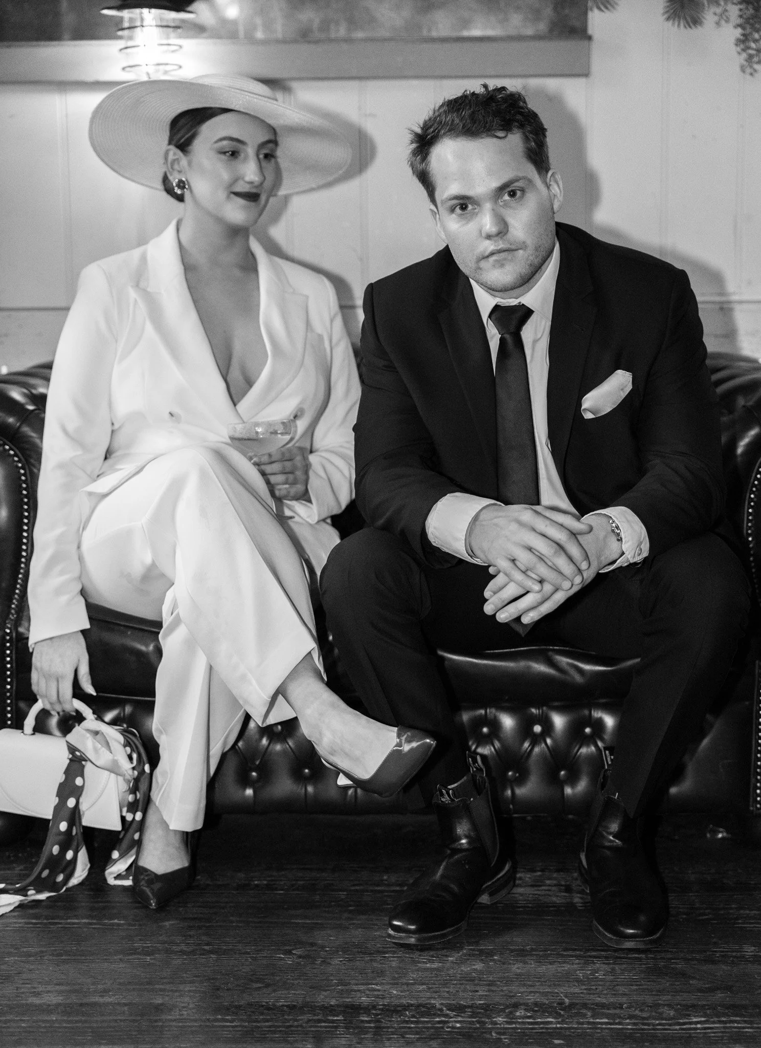 Black and white photo of a stylish woman in a wide-brimmed hat and a man in a suit sitting on a leather tufted bench in an indoor setting.