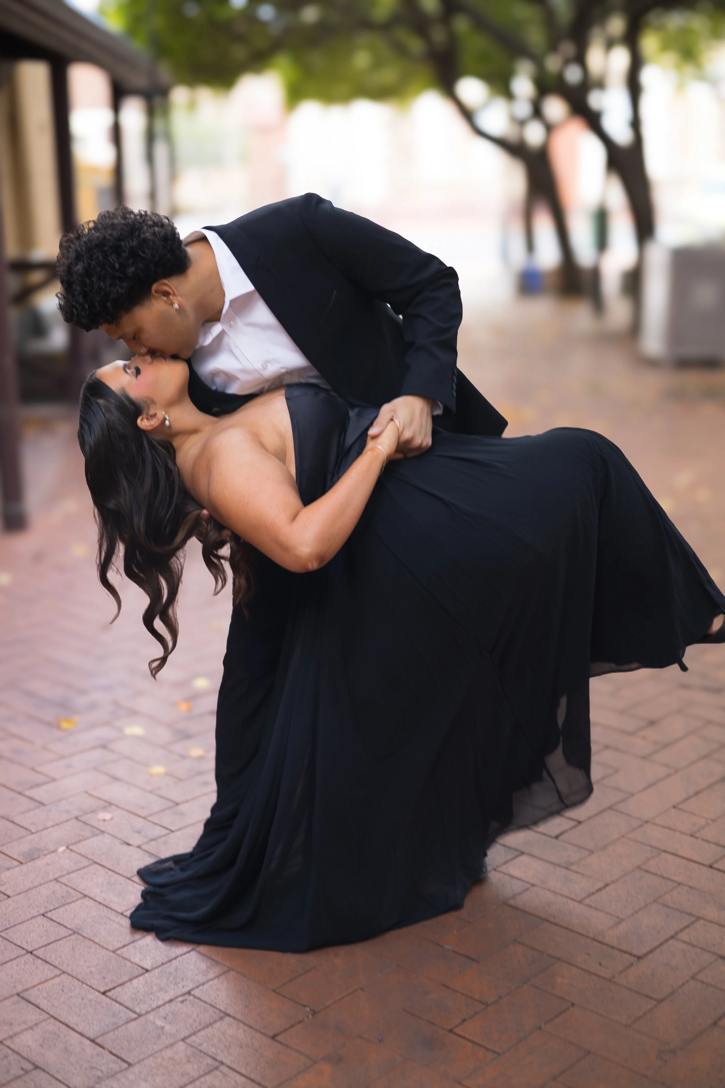 A couple dressed in formal attire dancing and sharing a kiss outdoors on a brick path, with trees and buildings in the background.