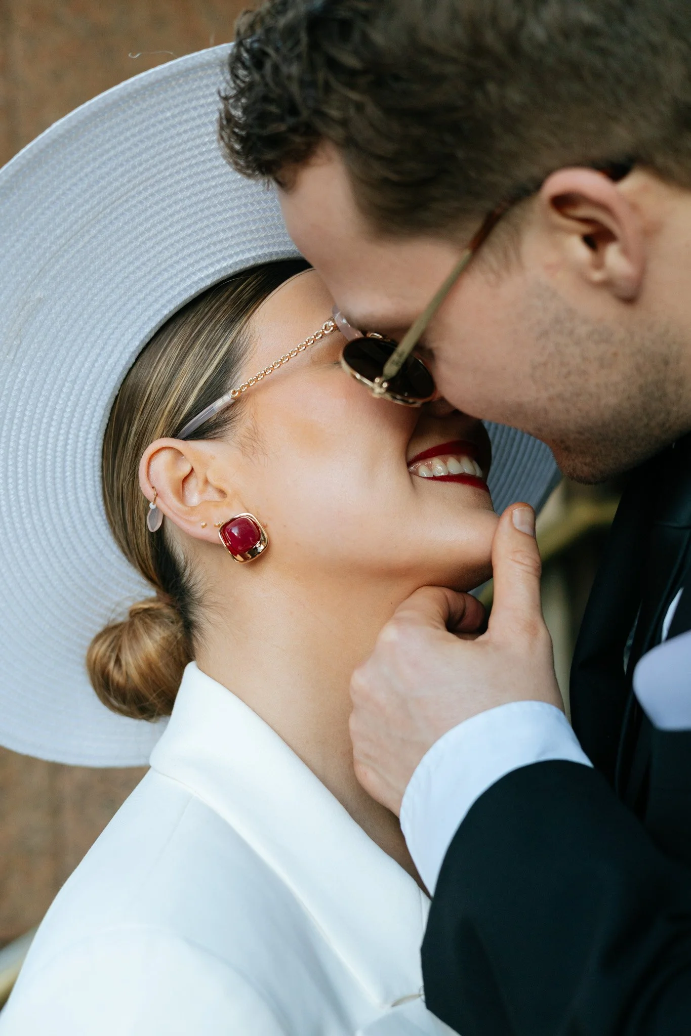 A couple dressed in formal attire sharing a kiss outdoors, woman wearing a wide-brimmed hat, earrings, necklace, and white outfit; man wearing glasses and a dark suit.