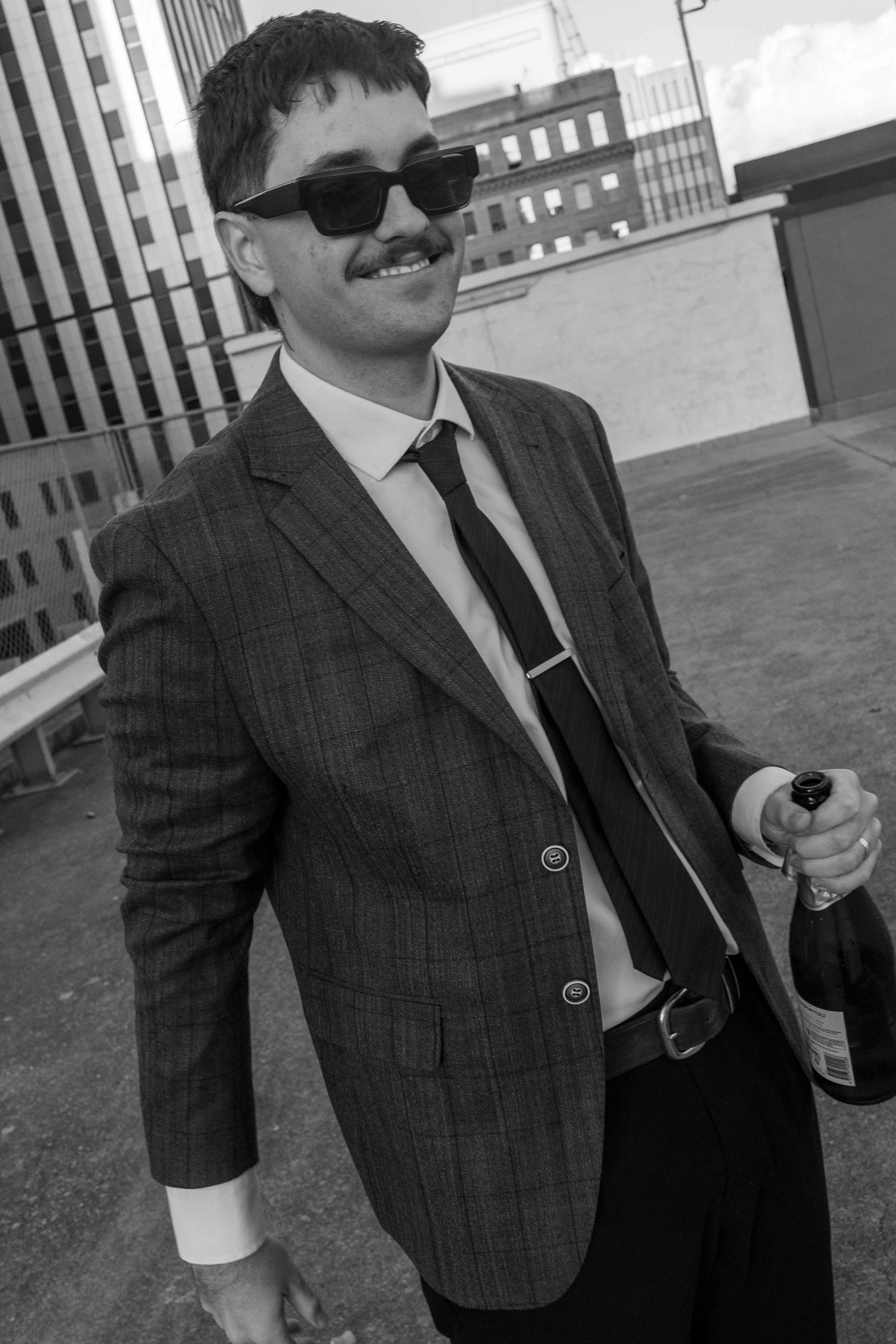 A man wearing sunglasses, a checkered blazer, and a white shirt with a tie, standing outdoors on a rooftop with a cityscape in the background, holding a bottle of champagne.