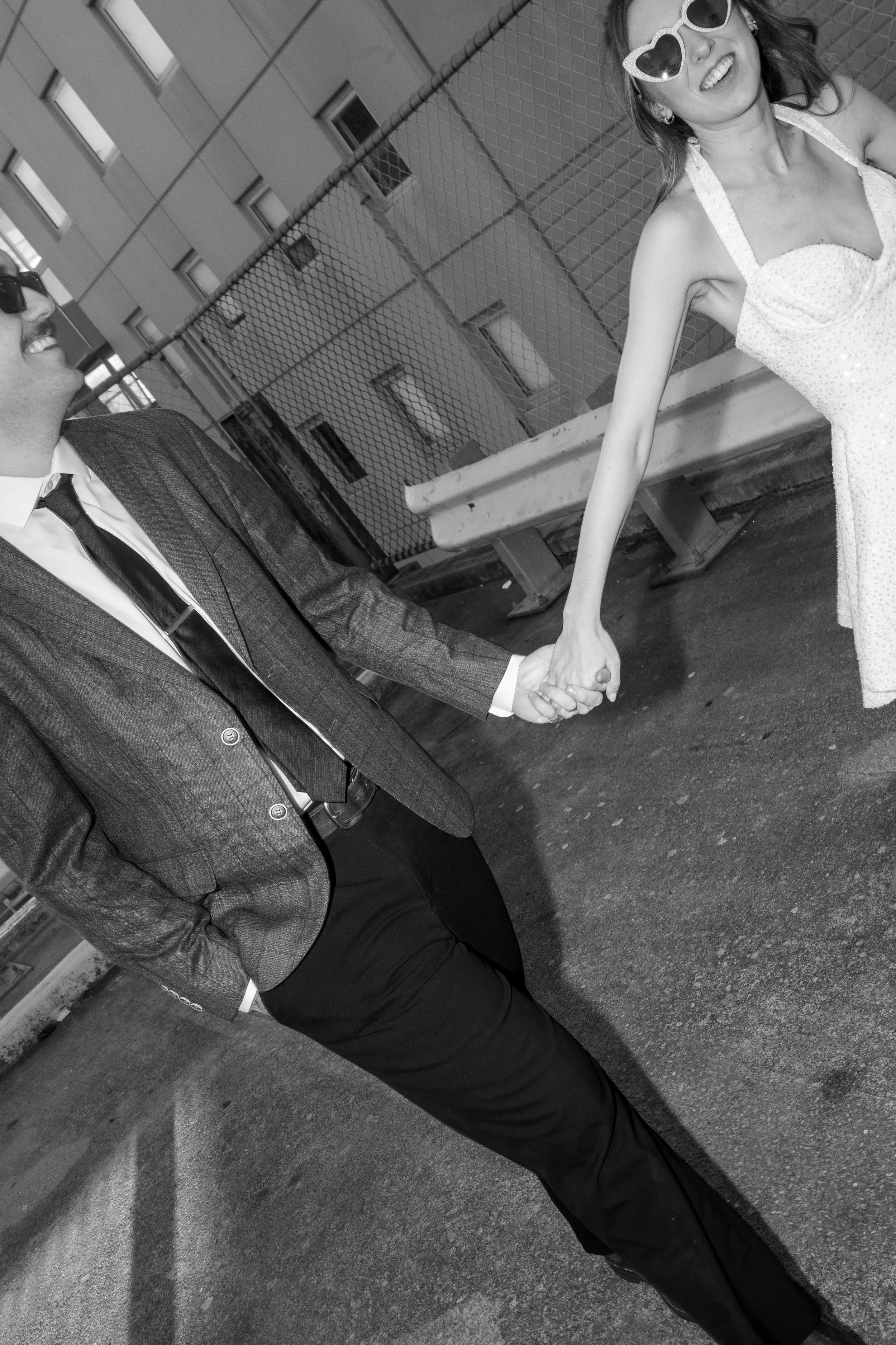 A woman wearing heart-shaped sunglasses and a sparkly dress is smiling and holding hands with a man dressed in a suit and tie, both standing outdoors near a building and a chain-link fence.