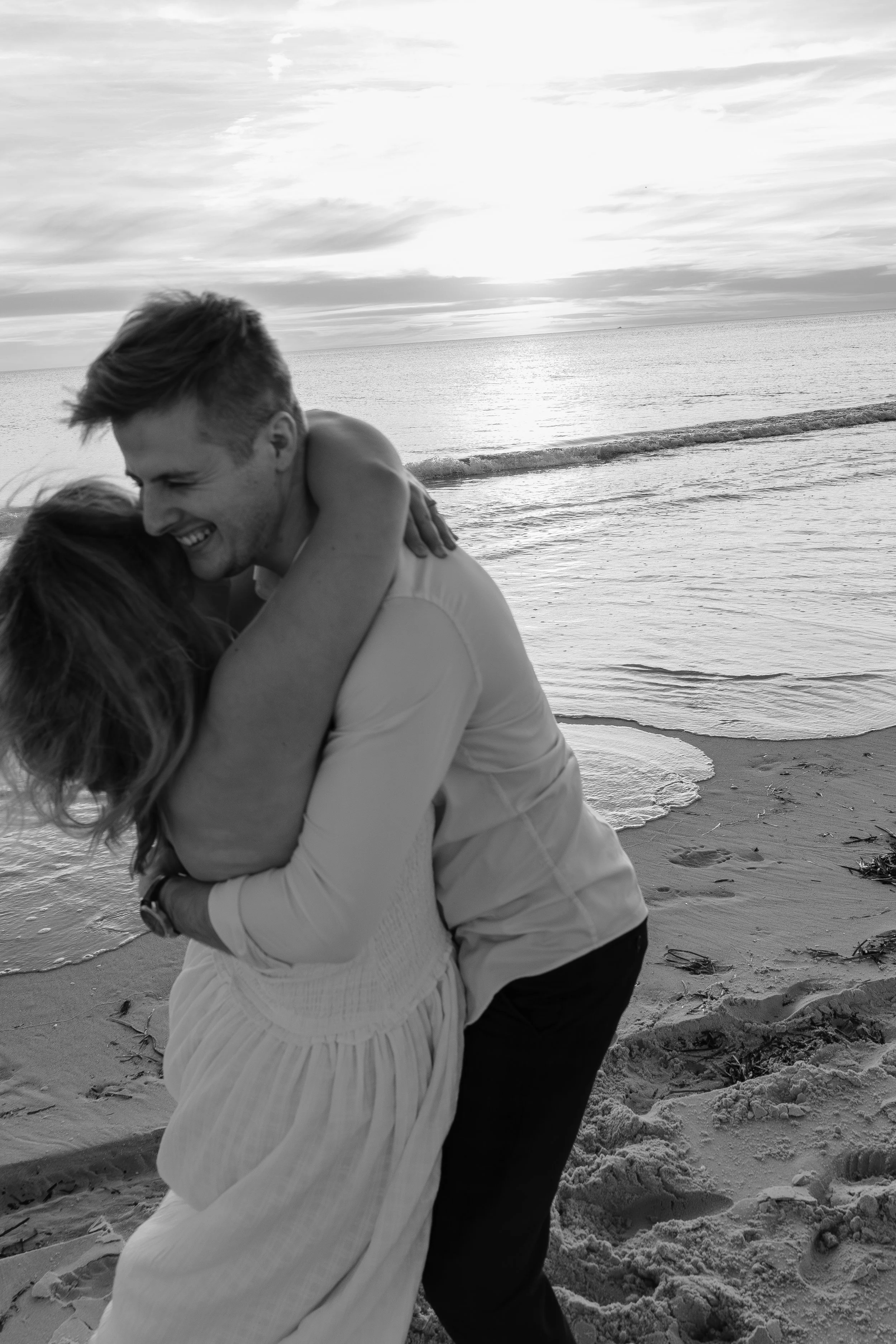 A couple embracing on the beach at sunset, with ocean waves and cloudy sky in the background.