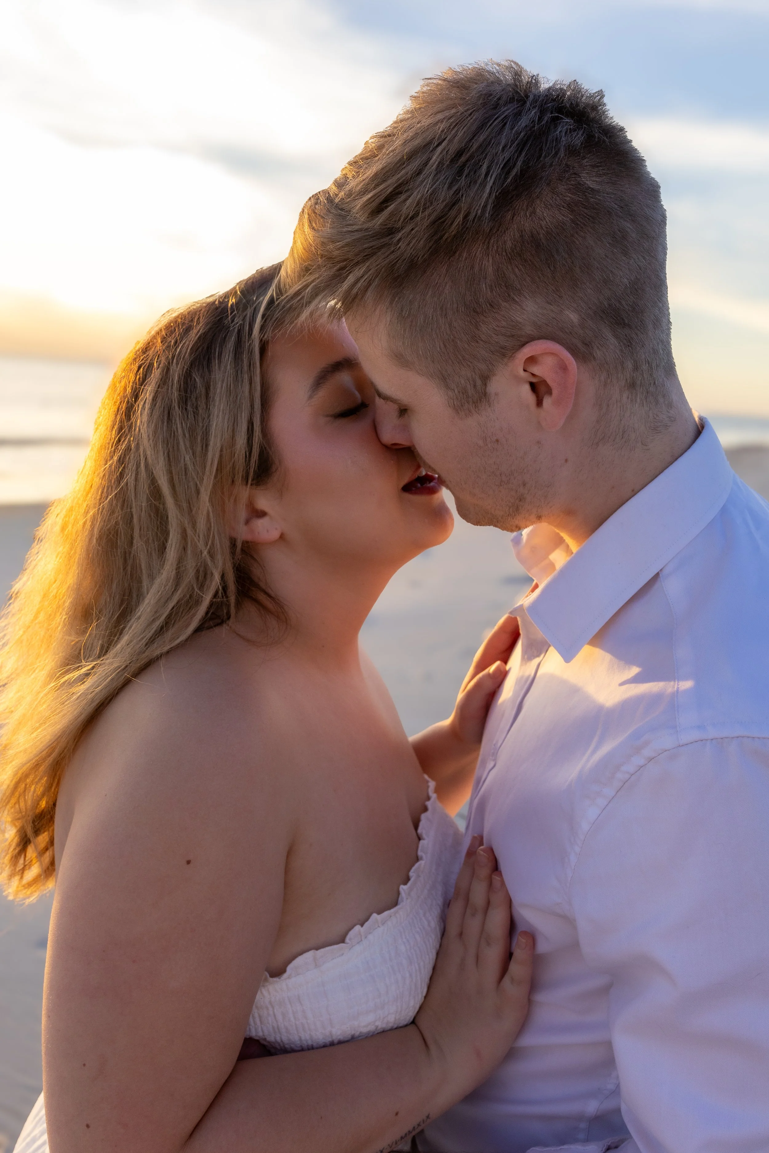 A couple kiss on the beach during sunset, with the ocean and sky in the background.