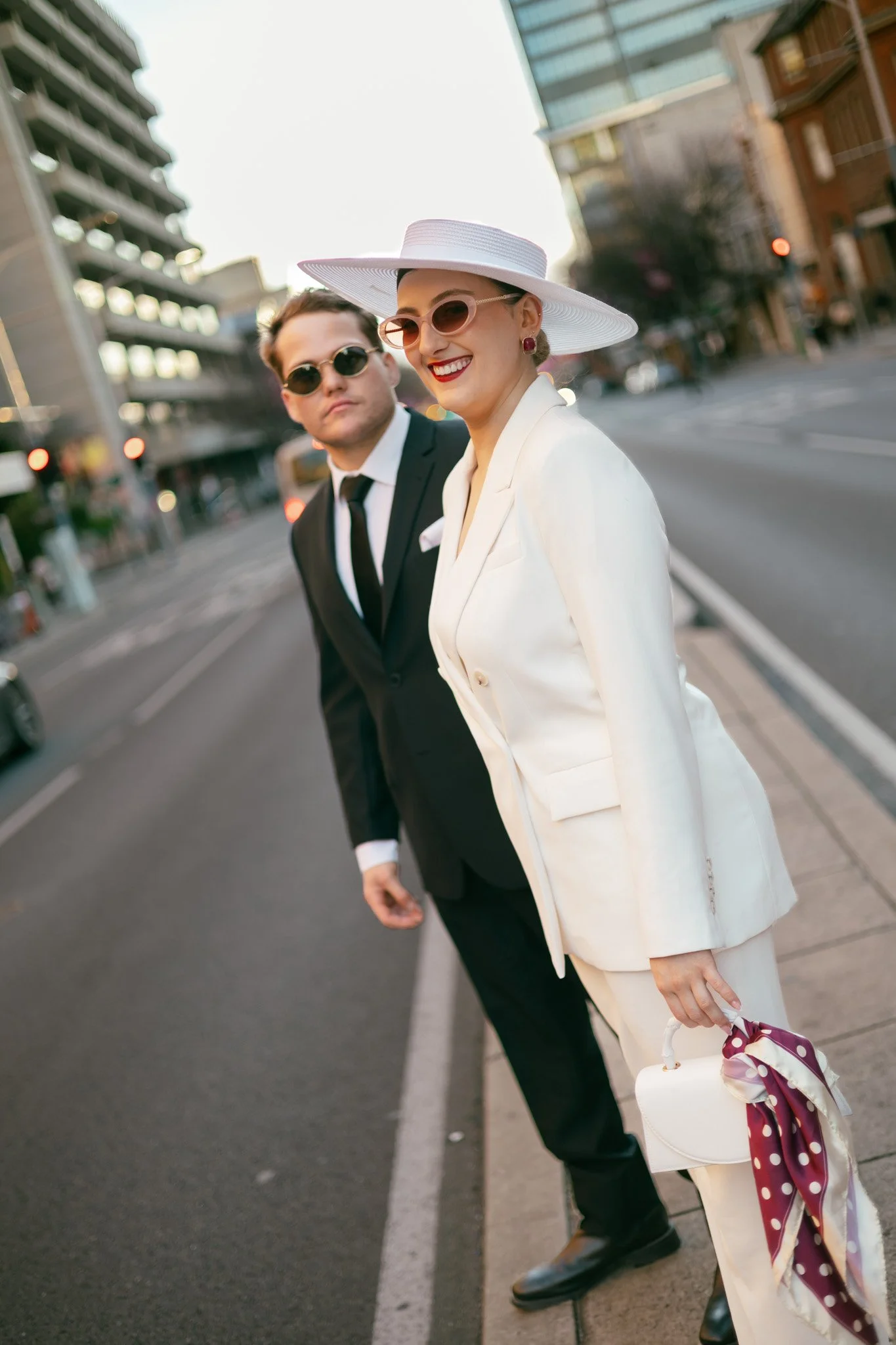 A woman and a man dressed in vintage style and sunglasses, standing on a city sidewalk. The woman wears a white wide-brimmed hat, a light-colored suit, and holds a small white handbag with a polka-dot scarf. The man wears a black suit, white shirt, a