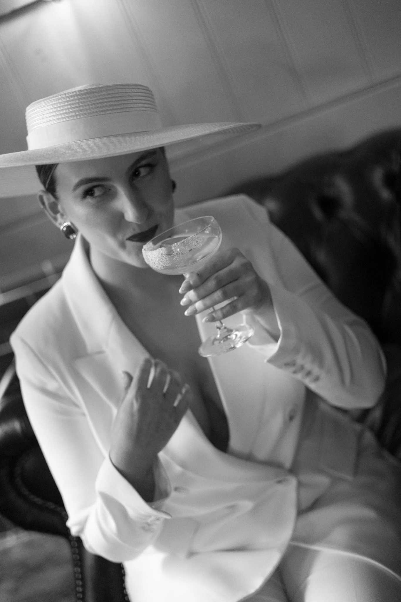 A woman wearing a wide-brimmed hat and dark lipstick, holding a cocktail glass with a sparkling drink, sitting on a leather chair in a dimly lit setting.