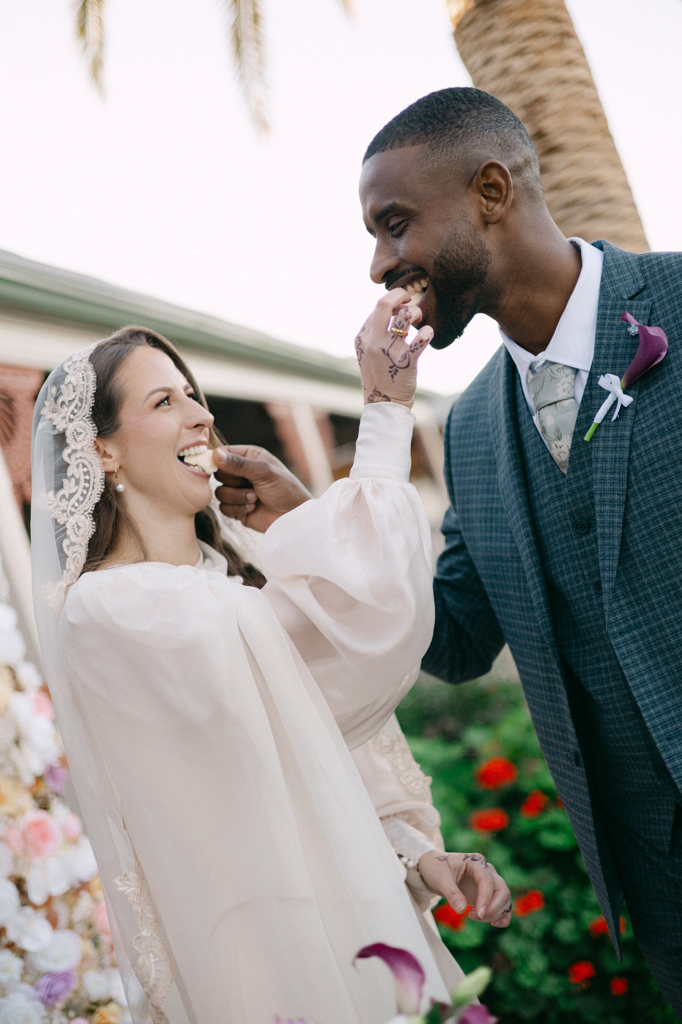 A bride and groom at a wedding ceremony, with a woman feeding the groom a piece of cake. The bride has a lace headband and white dress, while the groom is in a gray suit with a boutonniere. They are smiling, outdoors with palm trees and flower decorations.